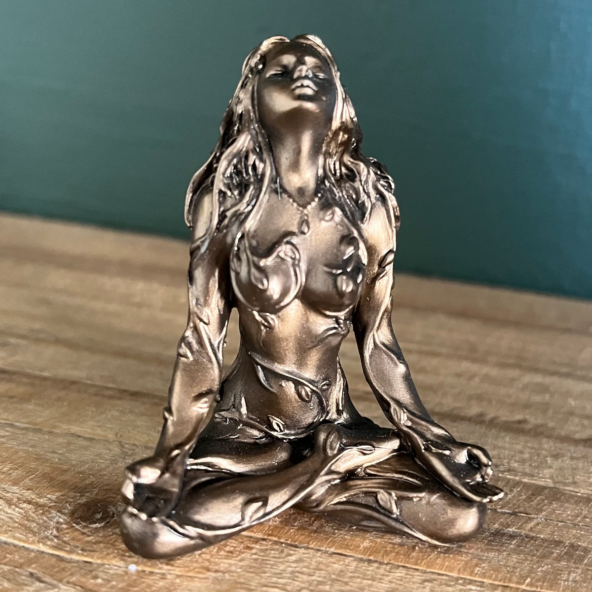 A metallic bronze figurine of a woman in a meditative pose, sitting on a wooden surface against a dark teal background.