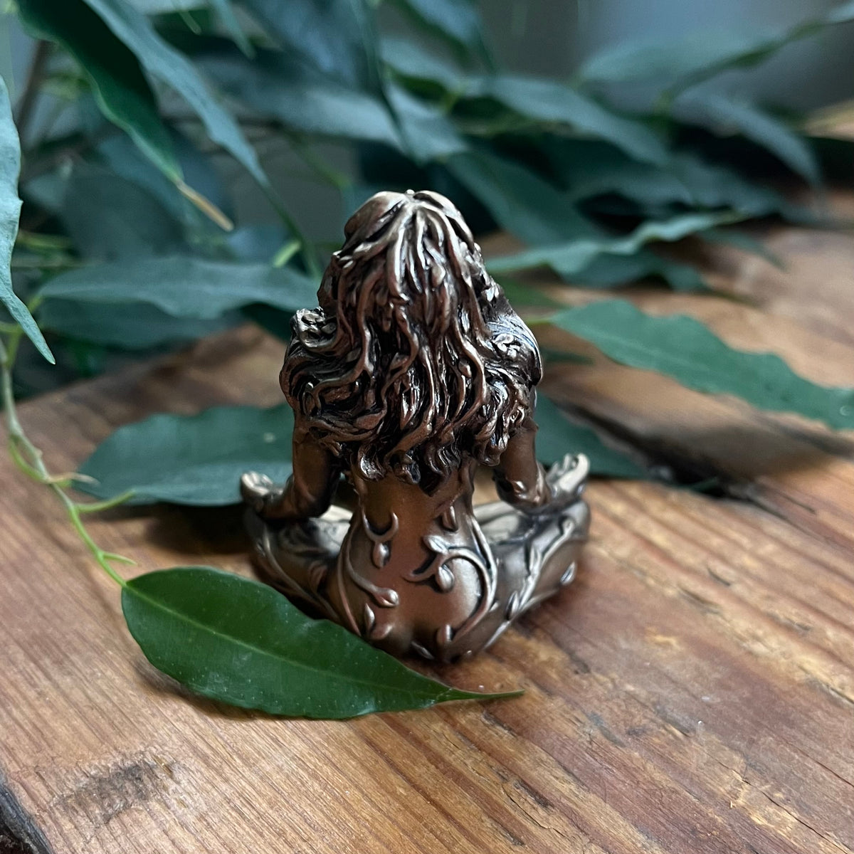 A small bronze-toned figurine of a seated figure with long wavy hair, photographed from the back on a wooden surface with green leaves around it.
