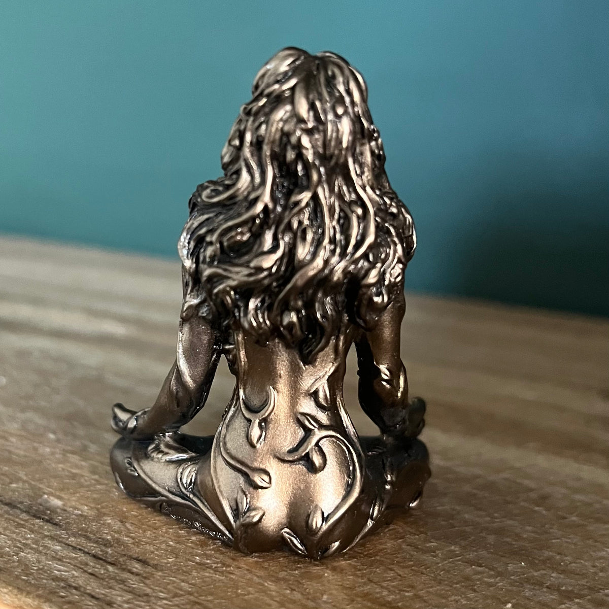 A small metallic figurine of a seated nude female figure with long wavy hair, shown from the back on a wooden surface against a teal-blue background.