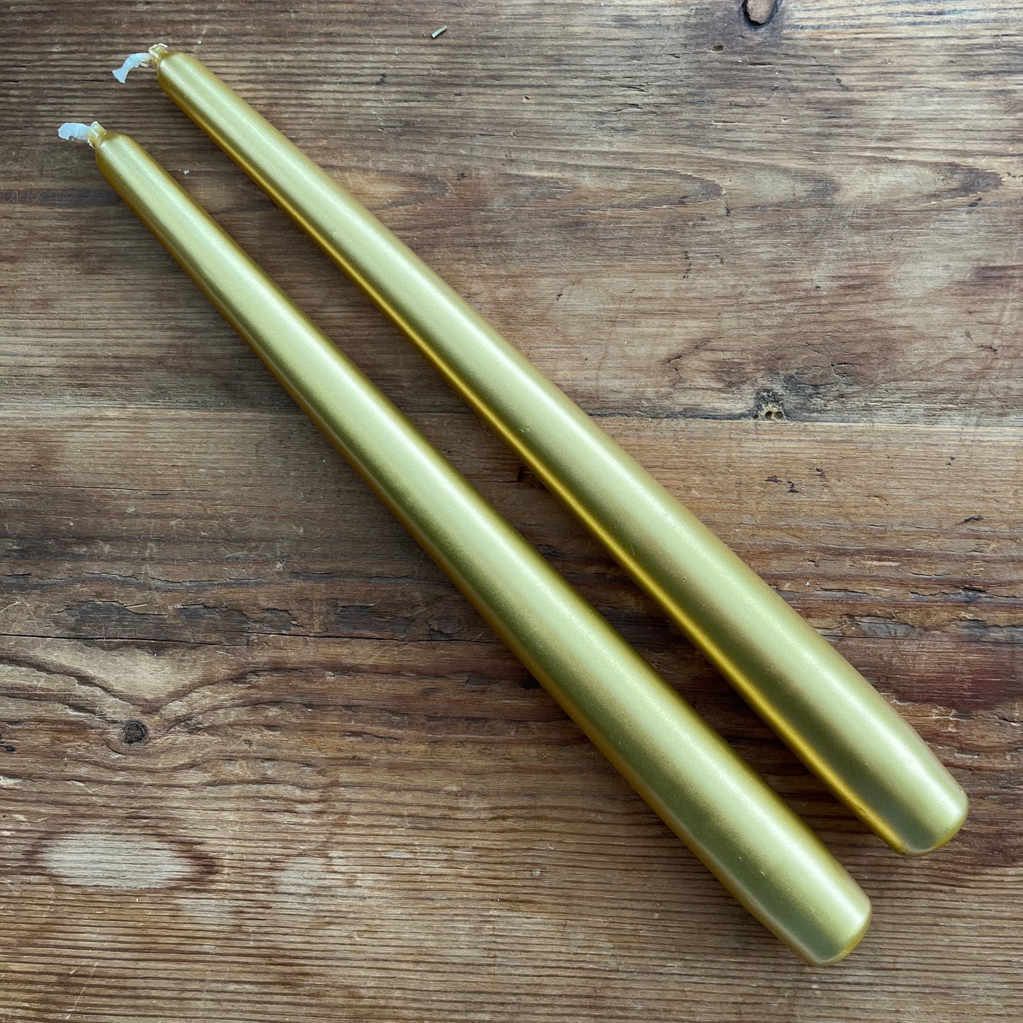 Two long tapered gold pillar candles with white wicks, placed diagonally on a rustic brown wooden surface.