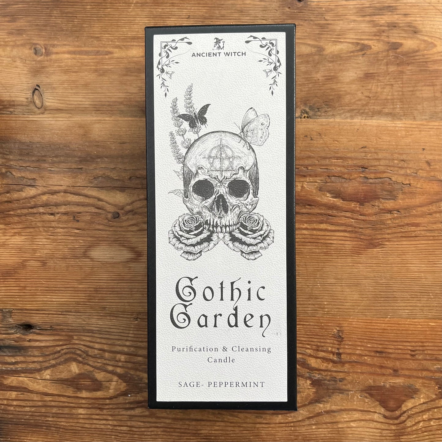 A framed rectangular candle box labeled 'Ancient Witch Gothic Garden Purification & Cleansing Candle' with a skull illustration, placed on a rustic wooden surface.