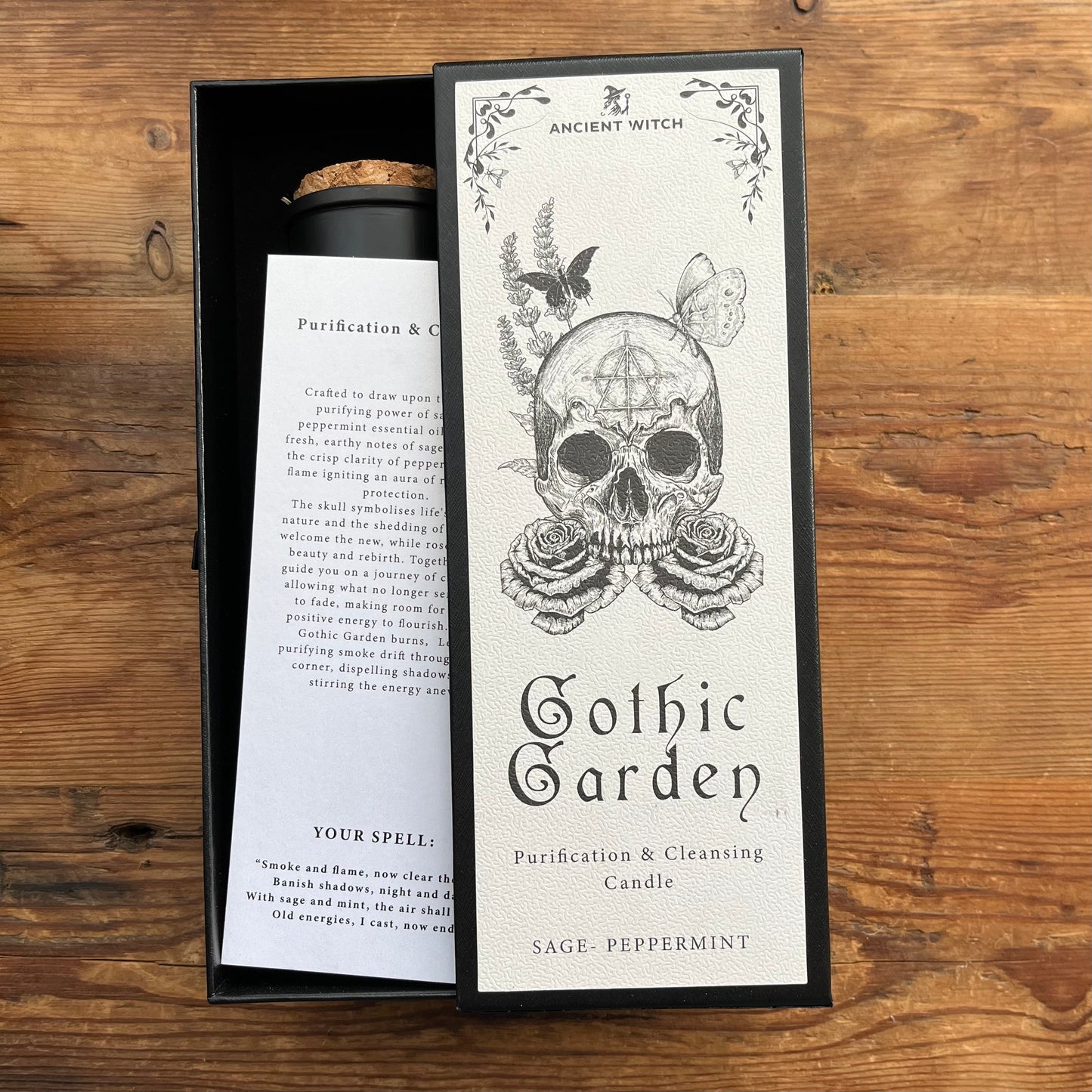 A boxed candle product labeled 'Ancient Witch Gothic Garden Purification & Cleansing Candle' in Sage-Peppermint scent, shown with its printed insert card beside the open black box on a wooden surface.