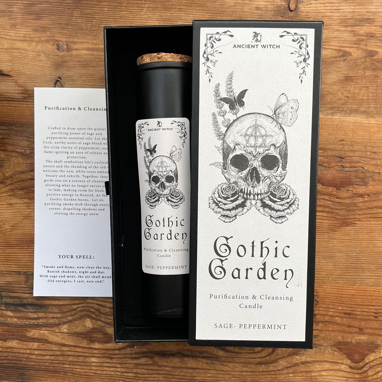 A black cylindrical candle jar with a cork stopper sits inside an open black presentation box next to its white front label and a printed instruction card, all placed on a wooden surface. The label reads 'Ancient Witch Gothic Garden Purification & Cleansing Candle' with a skull and rose illustration.