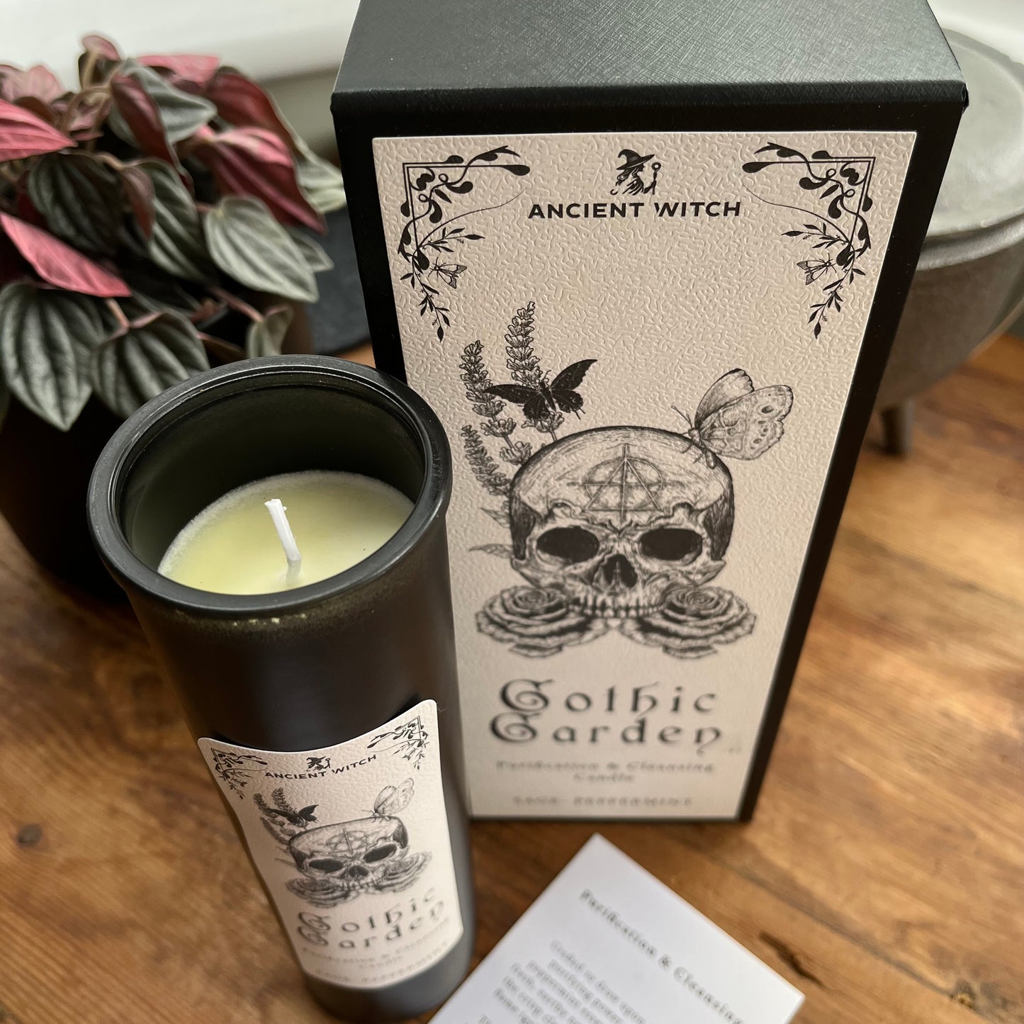A black glass candle jar with a white wax wick sits beside its matching black and cream packaging box on a wooden surface. The label reads 'ANCIENT WITCH Gothic Gardep' with a skull, butterfly, and floral illustration.