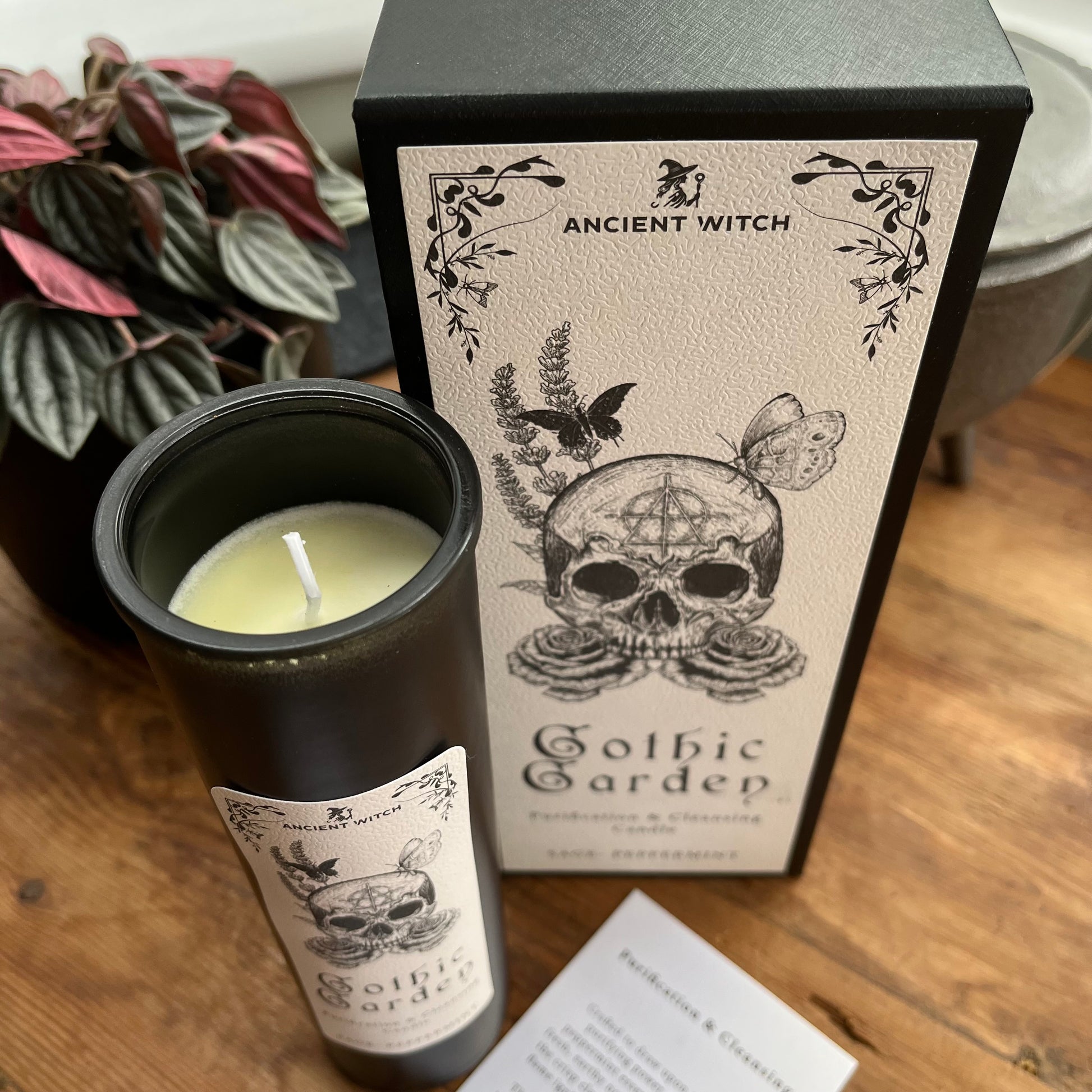 A black glass candle jar with a white wax wick sits beside its matching black and cream packaging box on a wooden surface. The label reads 'ANCIENT WITCH Gothic Gardep' with a skull, butterfly, and floral illustration.
