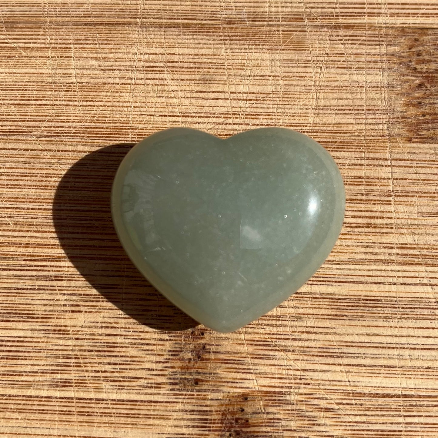 A polished heart-shaped stone in a muted sage green color, resting on a textured wooden surface.