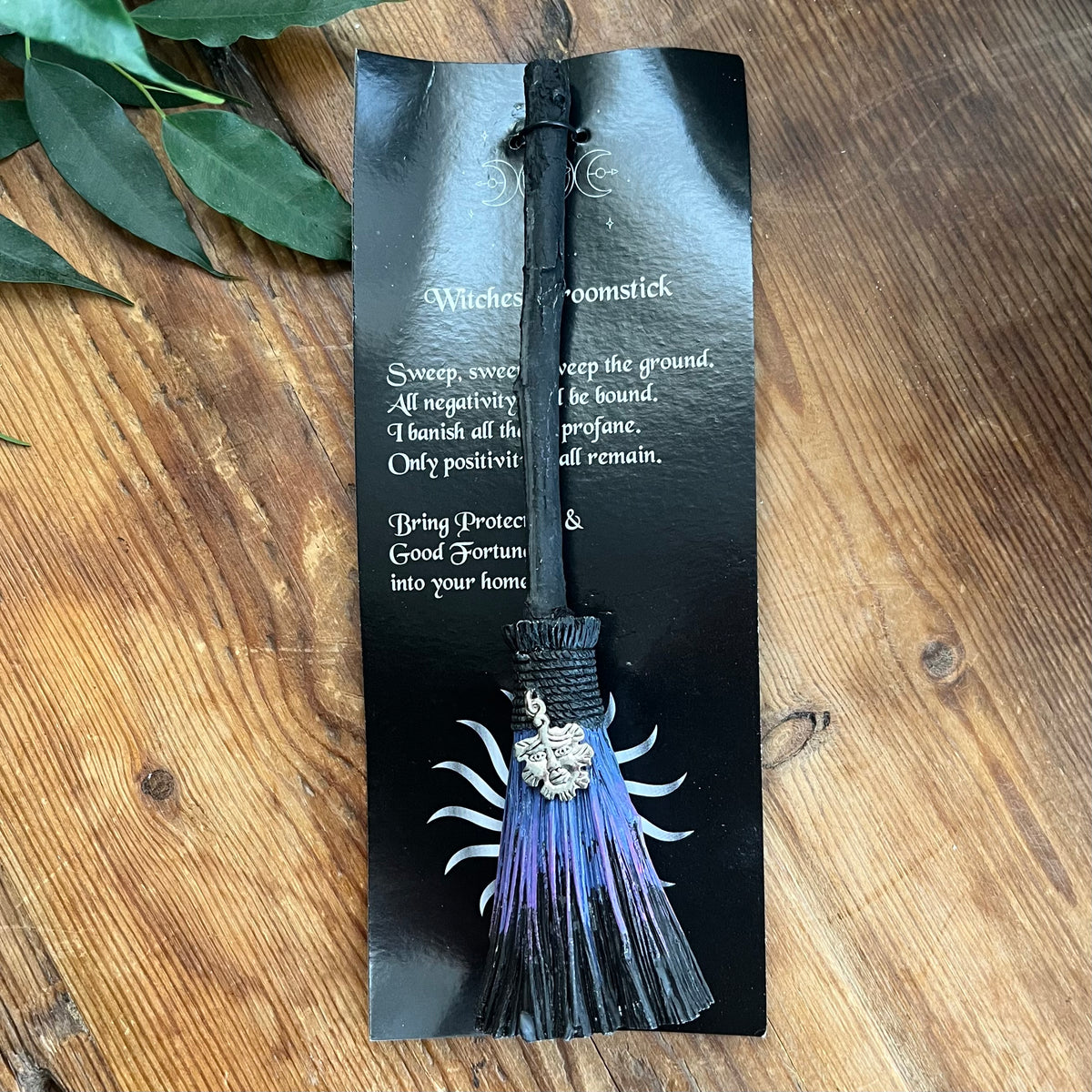 A black witch's broomstick tassel bookmark or tag with a silver moon charm, resting on its black backing card over a wooden surface.
