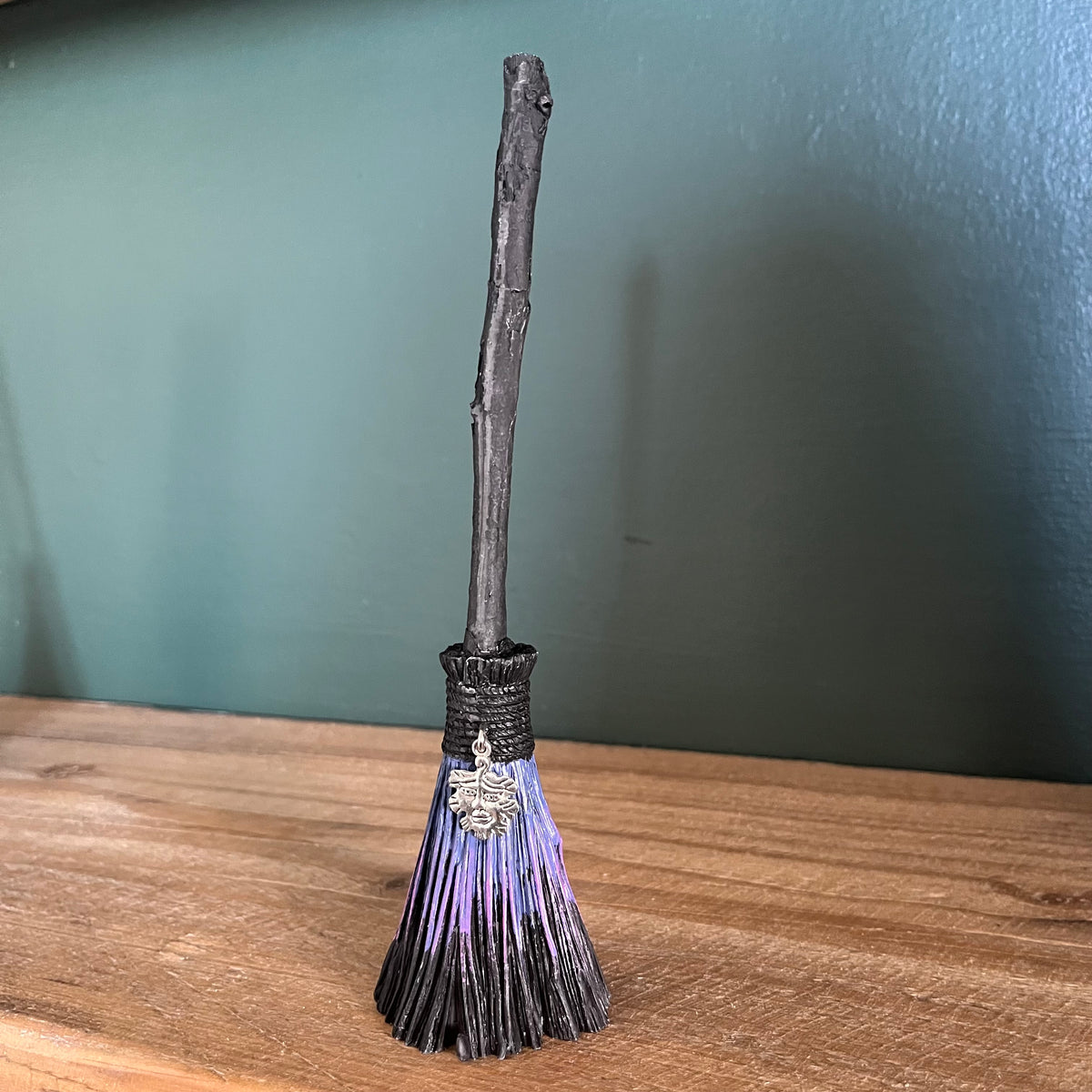 A miniature witch's broom with a dark wooden handle, black bristles, and purple tips, featuring a silver charm, set on a wooden surface against a dark teal wall.