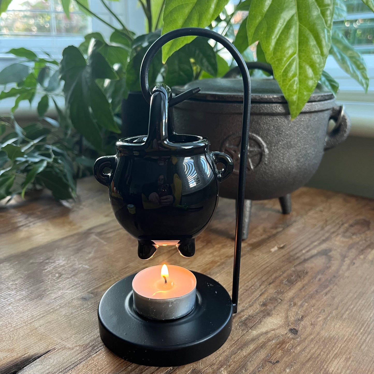 Black ceramic oil burner with a lit candle on a wooden surface, surrounded by plants.
