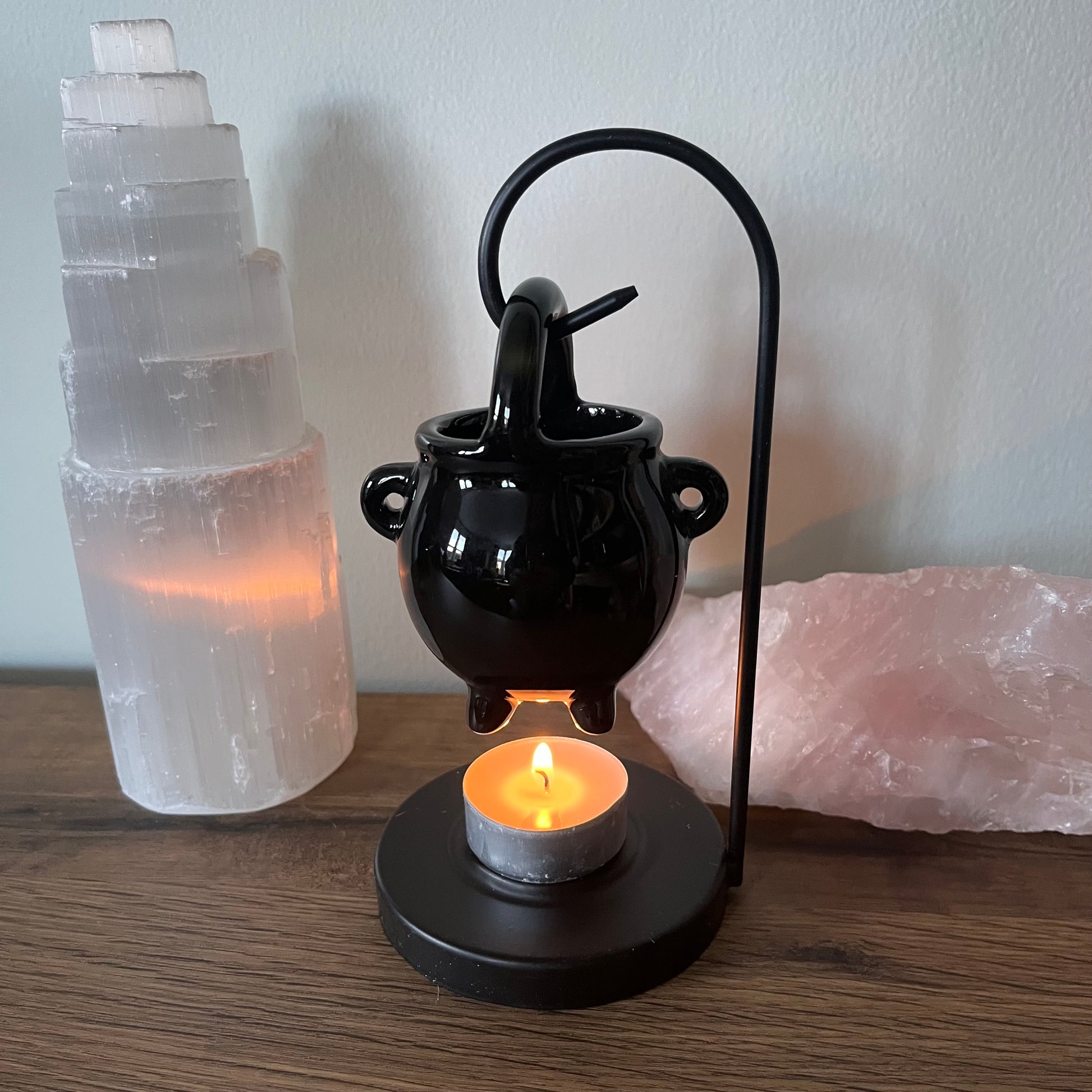 Black ceramic cauldron shaped oil burner with a lit candle, surrounded by crystals on a wooden surface.