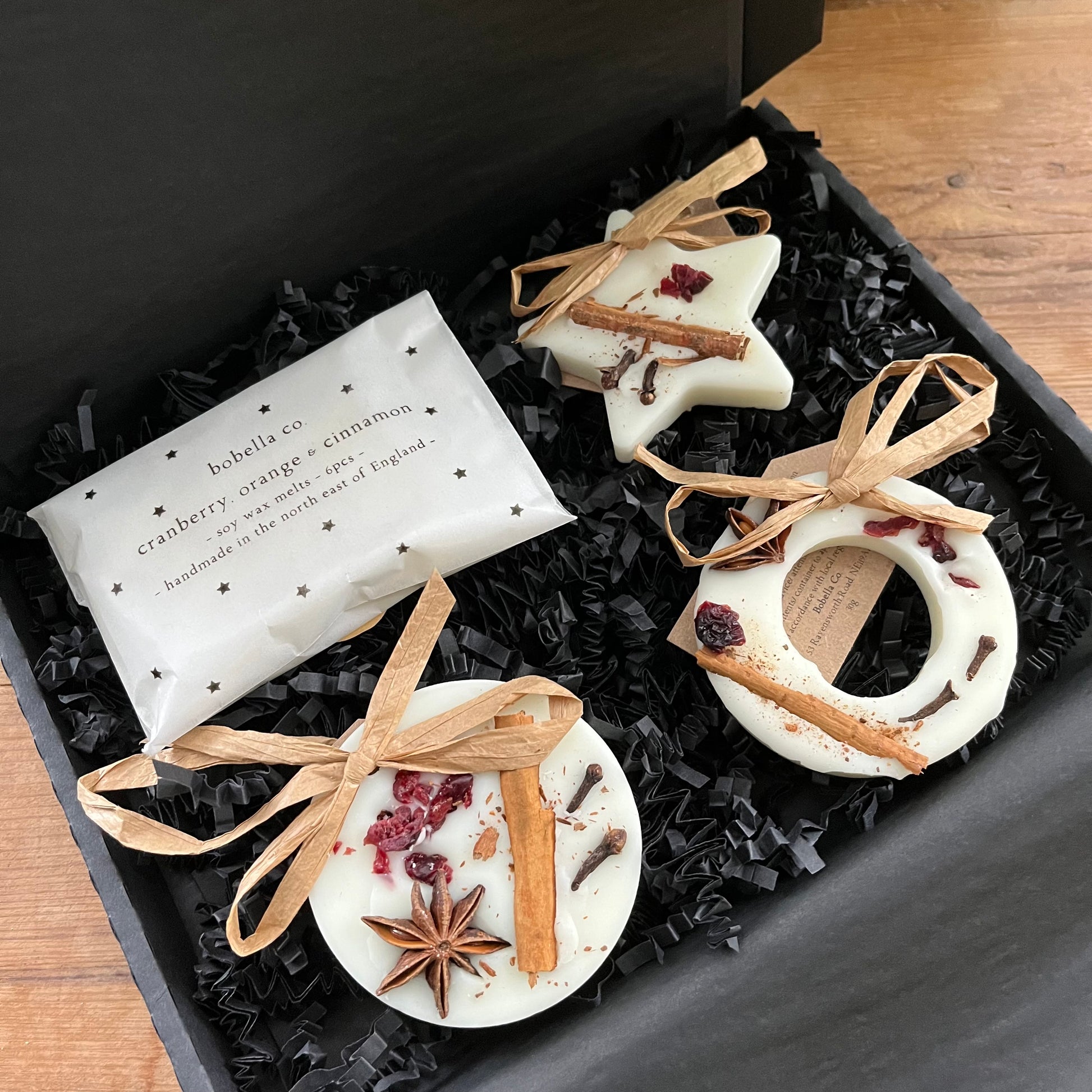 Three festive shaped hanging wax melt decorations and a pack of mini wax melts n a gift box,  on a wooden surface.  