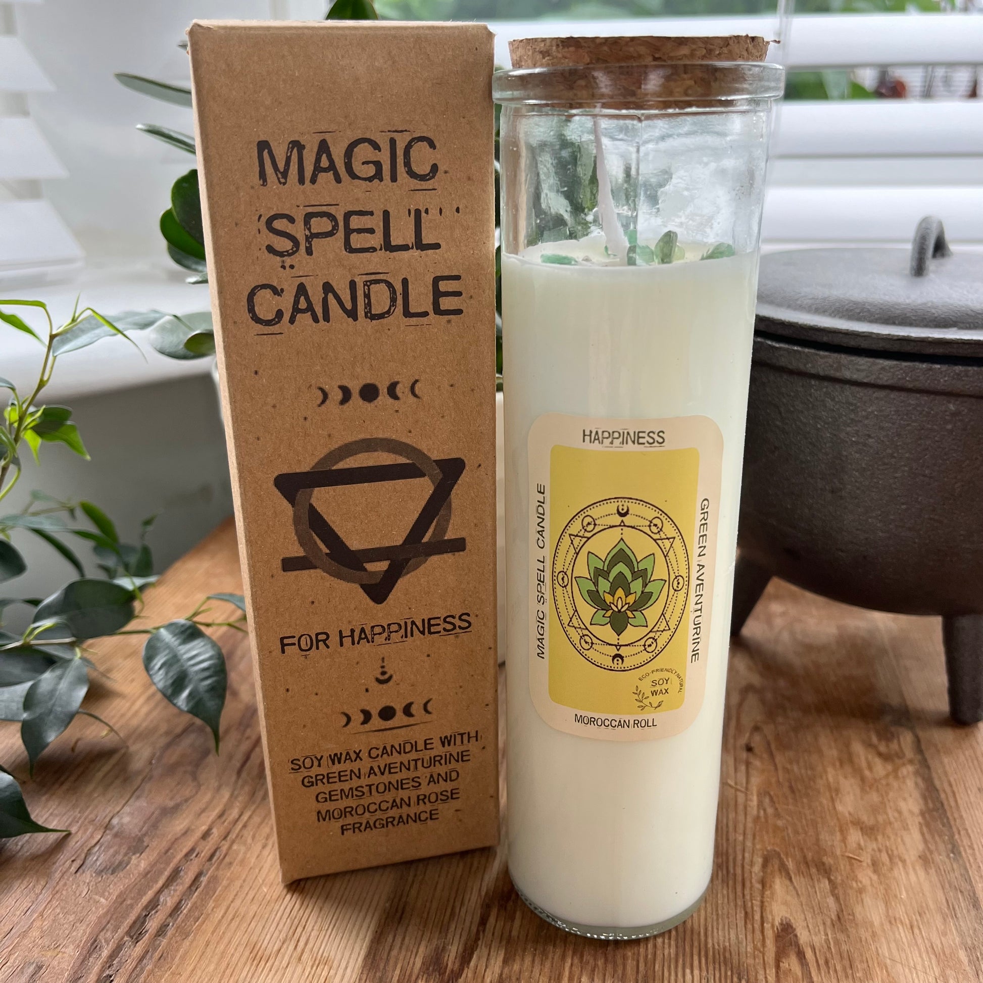 Tall glass spell candle with cork lid. The label on the candle features an image of a Lotus Leaf and the words Happiness, Green Aventurine and Moroccan Rose . The candle itself is white with Green Aventurine gemstones embedded within it. Alongside the glass Magic spell candle is it's brown cardboard box which has the words magic spell candle for Happiness, soy wax candle with Green Aventurine gemstones and Moroccan Rose fragrance.