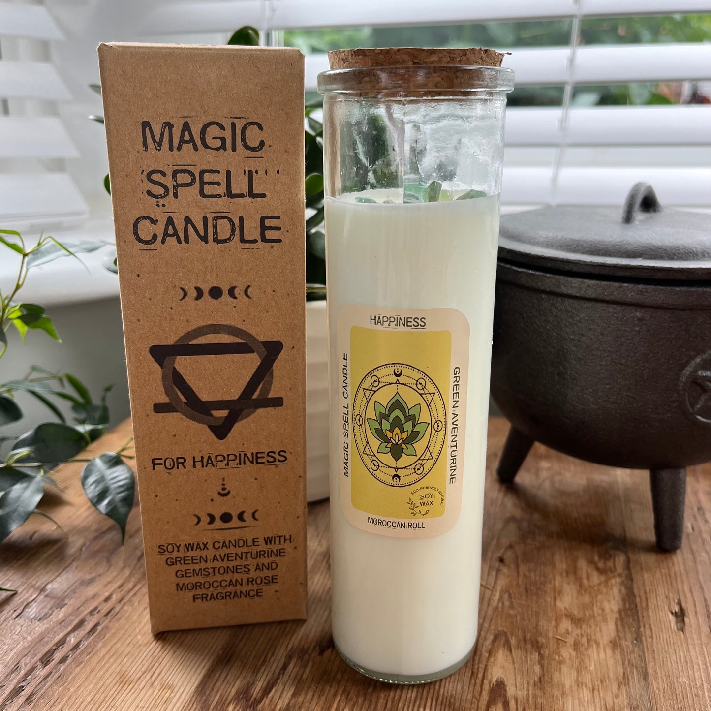 Tall glass spell candle with cork lid. The label on the candle features an image of a Lotus Leaf and the words Happiness, Green Aventurine and Moroccan Rose . The candle itself is white with Green Aventurine gemstones embedded within it. Alongside the glass Magic spell candle is it's brown cardboard box which has the words magic spell candle for Happiness, soy wax candle with Green Aventurine gemstones and Moroccan Rose fragrance.