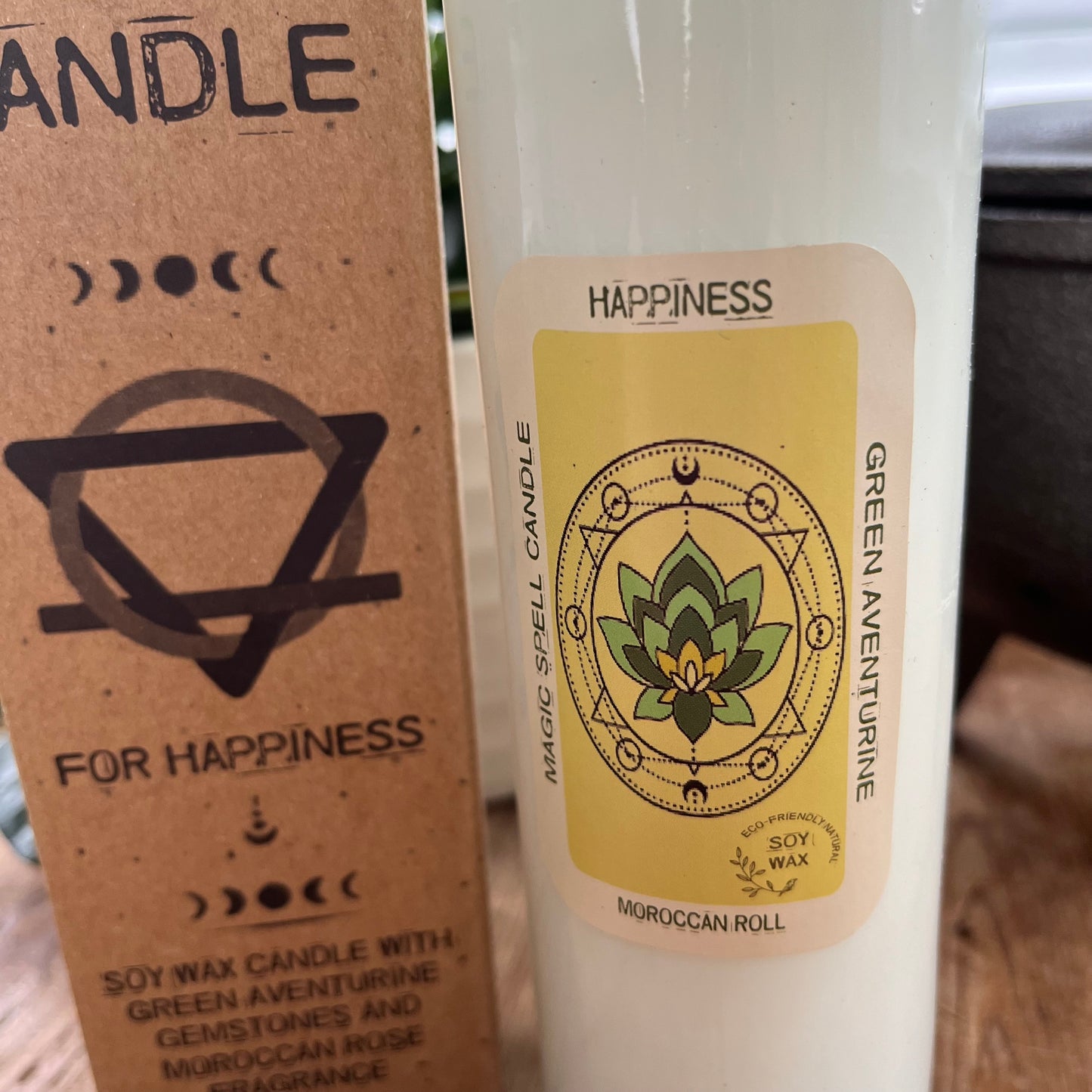 Tall glass spell candle with cork lid. The label on the candle features an image of a Lotus Leaf and the words Happiness, Green Aventurine and Moroccan Rose . The candle itself is white with Green Aventurine gemstones embedded within it. Alongside the glass Magic spell candle is it's brown cardboard box which has the words magic spell candle for Happiness, soy wax candle with Green Aventurine gemstones and Moroccan Rose fragrance.