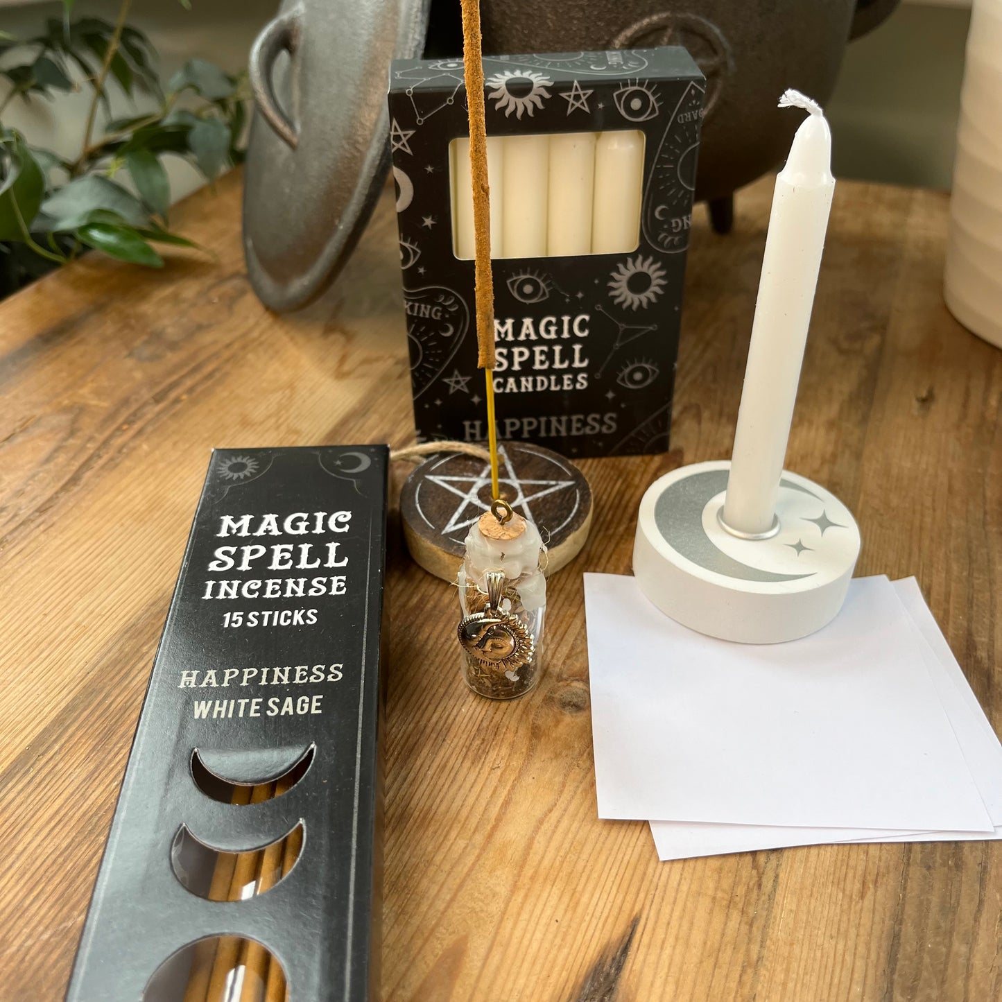 'Happiness Spell Kit' Contents displayed on a wooden table. Pack of Happiness white sage Incense sticks, and wooden pentagram incense holder, Pack of white magic spell candles, wooden white spell candle holder with crescent moon and star design, squares of white paper and a happiness spell bottle containing herbs and sealed with white wax and a charm.