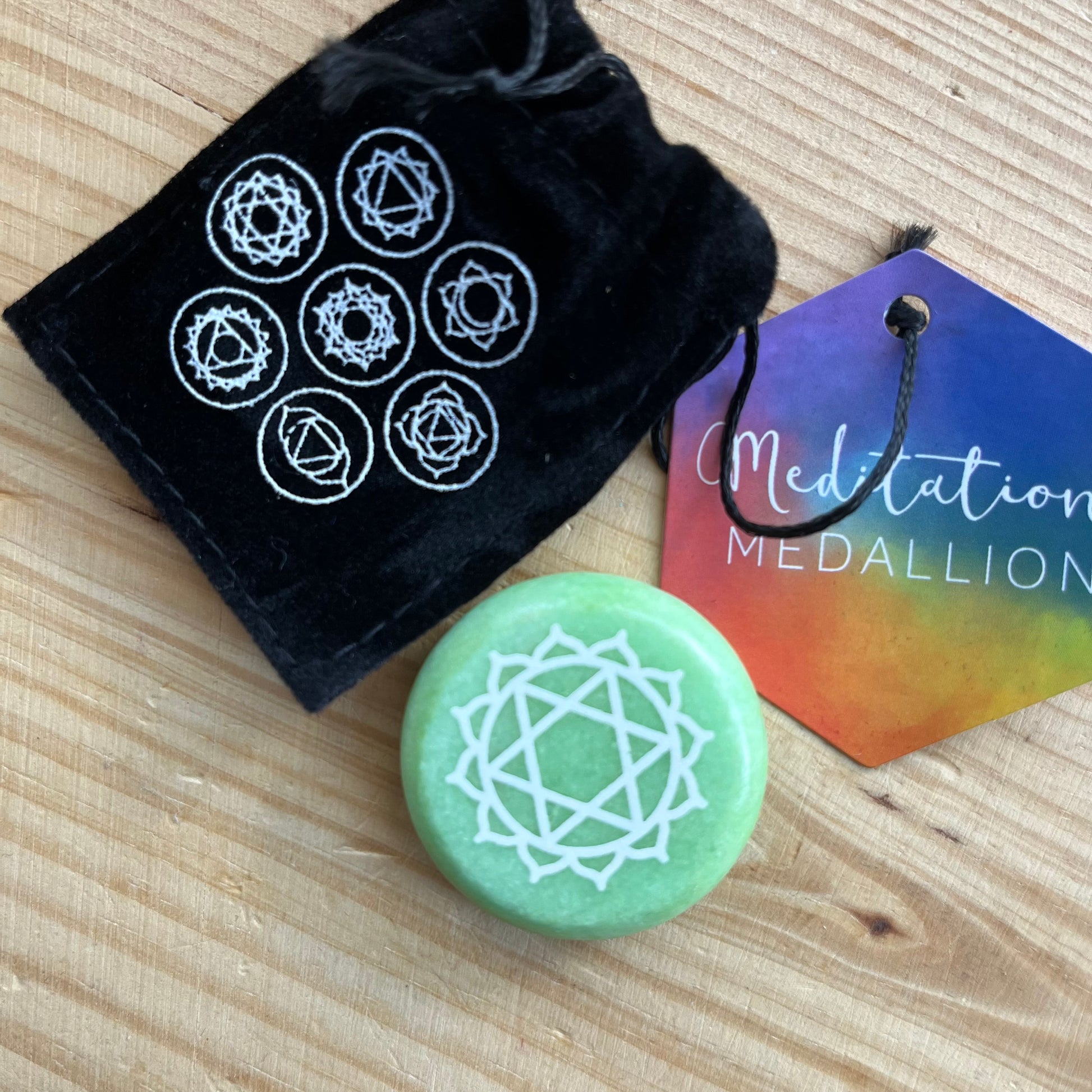 Green circular flat meditation stone is engraved with the Heart Chakra symbol alongside it's black velvet pouch printed with the 7 Chakra sybmols 