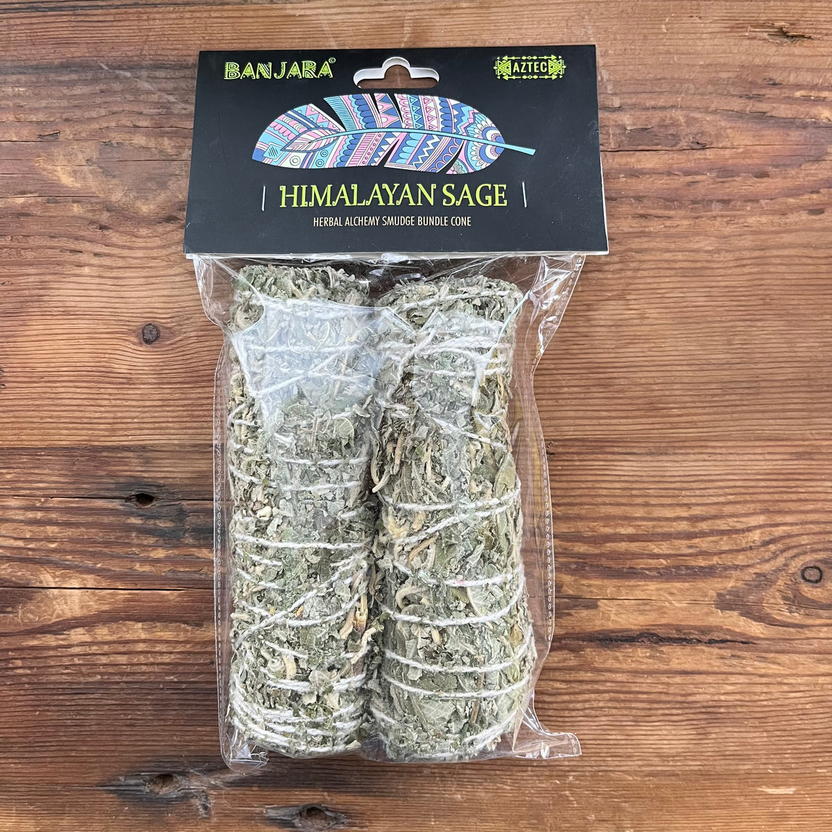 Two bundled sticks of Himalayan sage smudge sticks in clear plastic packaging with a black header card, placed on a wooden surface.