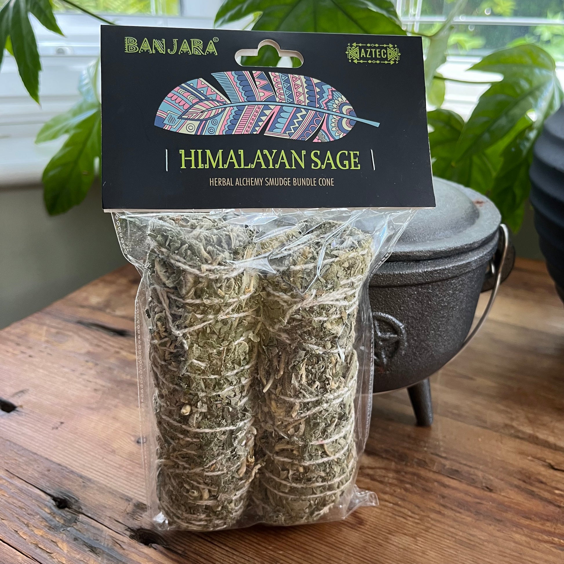 A packaged bundle of two sage smudge cones in a clear plastic bag with a black header card reading 'BANJARA' and 'HIMALAYAN SAGE'. The product sits on a wooden table with a cast iron pot and green plants in the background.