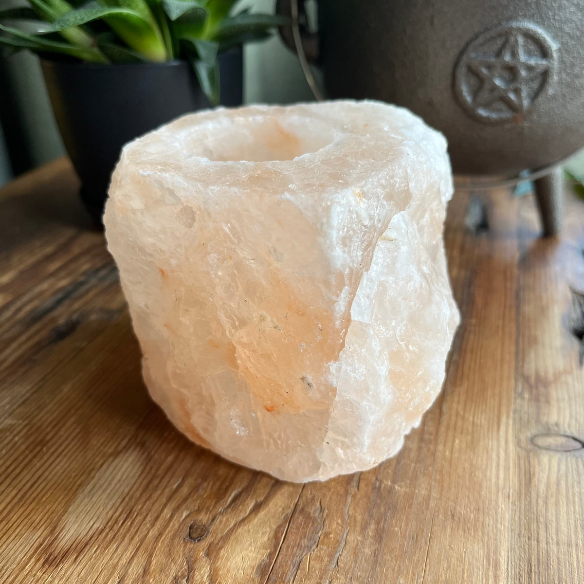 A raw, rough-cut salt crystal candle holder with a hollow center, resting on a wooden tabletop with a potted plant and a decorative metal vessel in the background.