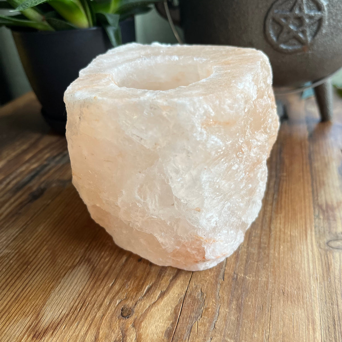 A raw, rough-cut pinkish-white salt stone candle holder with a central hole, resting on a wooden tabletop with potted plants and a cast iron pot blurred in the background.