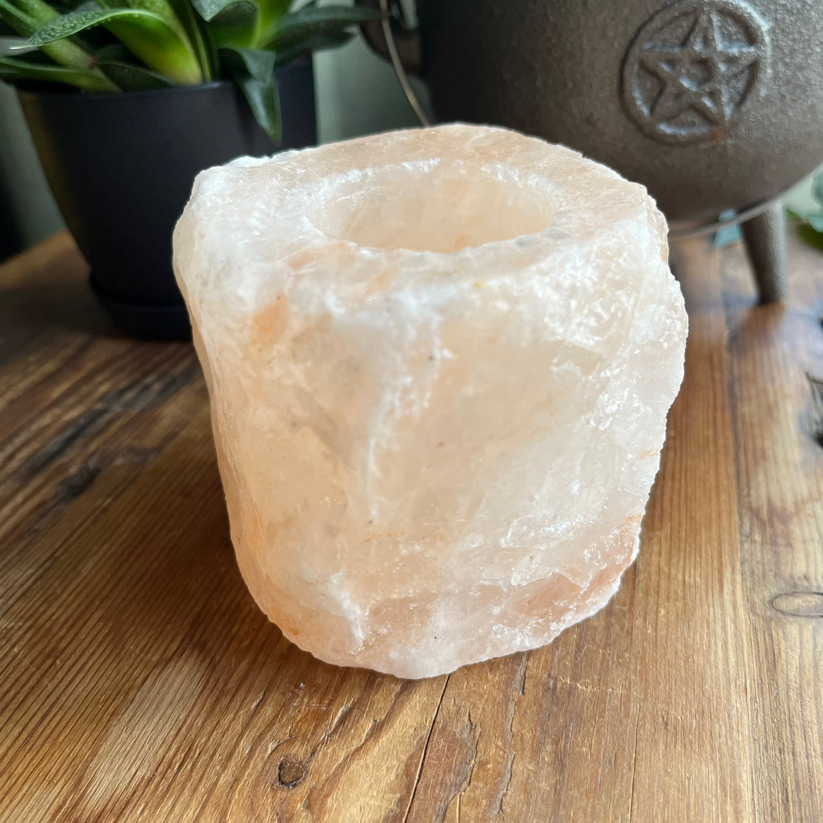 A raw, rough salt candle holder made from a natural pinkish-white salt stone, placed on a wooden tabletop with a potted plant and a cast iron pot in the background.