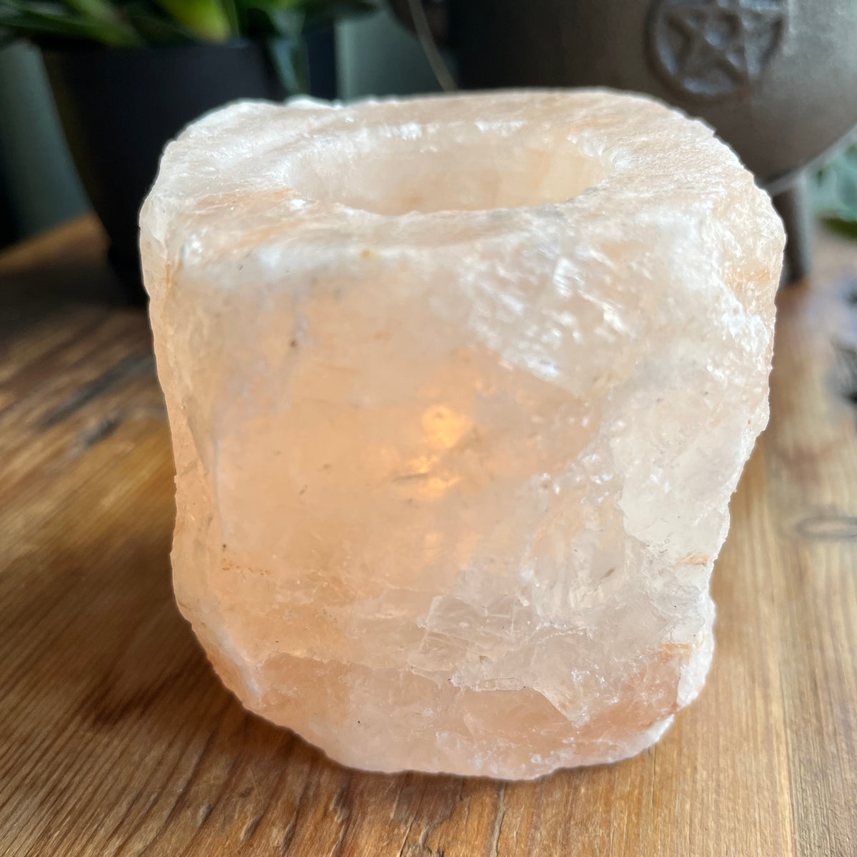 A raw, unpolished pinkish-white salt crystal Tealight holder sitting on a wooden tabletop, glowing with a warm light from within.