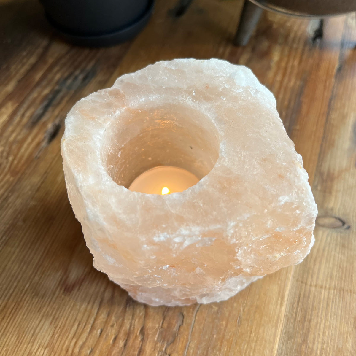 A raw pink salt crystal candle holder with a circular hole in the center, containing a lit tea light candle, placed on a wooden tabletop.