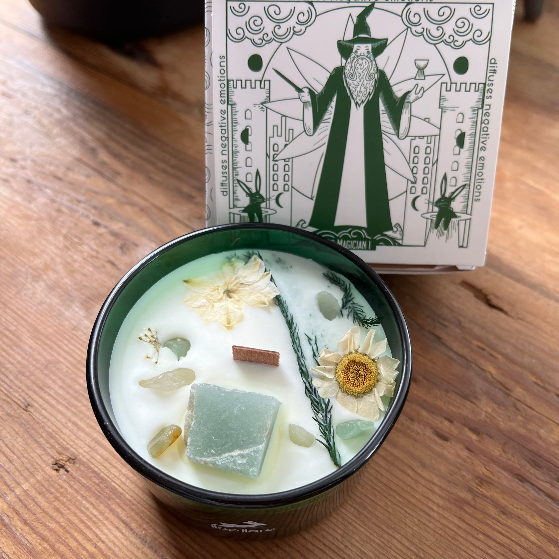 Candle in a Green container with floral elements on a wooden surface, accompanied by a decorative Magician tarot card.