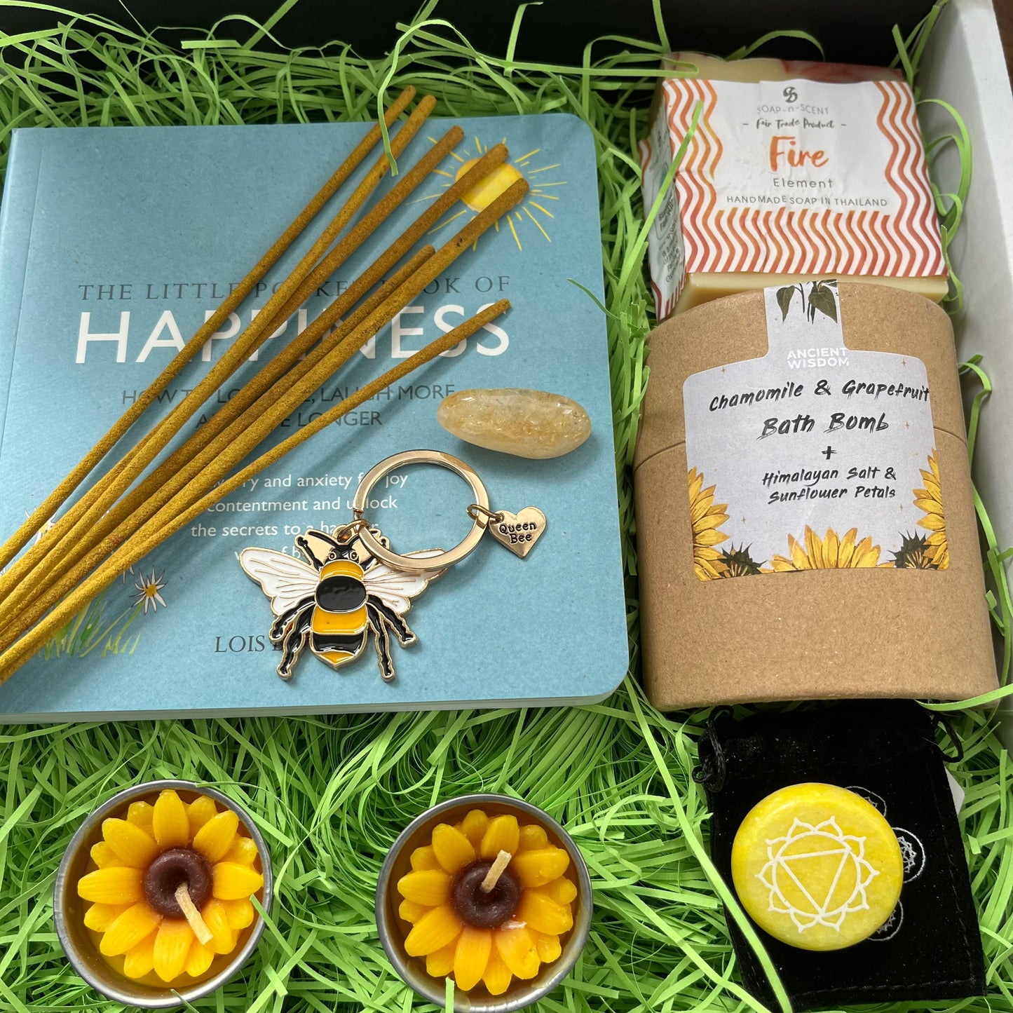 A curated gift set arranged in green shredded paper inside a box, featuring a light blue book, wooden incense sticks, a bee-shaped keychain, a bath bomb labeled chamomile & grapefruit, two yellow flower-shaped tea lights, and a yellow gemstone on a black background.