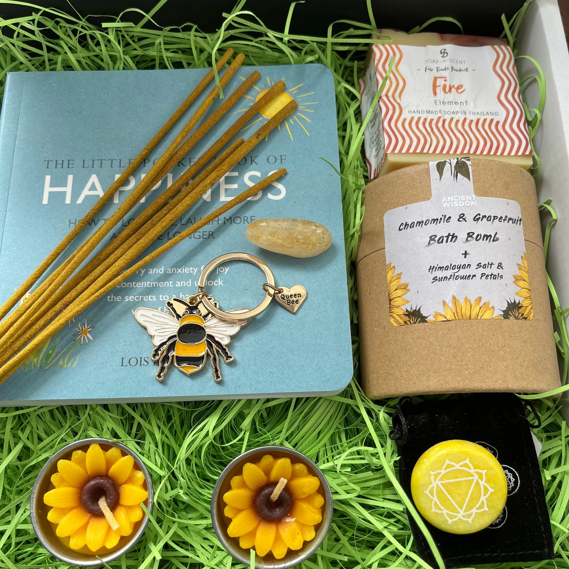 A curated gift set arranged in green shredded paper inside a box, featuring a light blue book, wooden incense sticks, a bee-shaped keychain, a bath bomb labeled chamomile & grapefruit, two yellow flower-shaped tea lights, and a yellow gemstone on a black background.