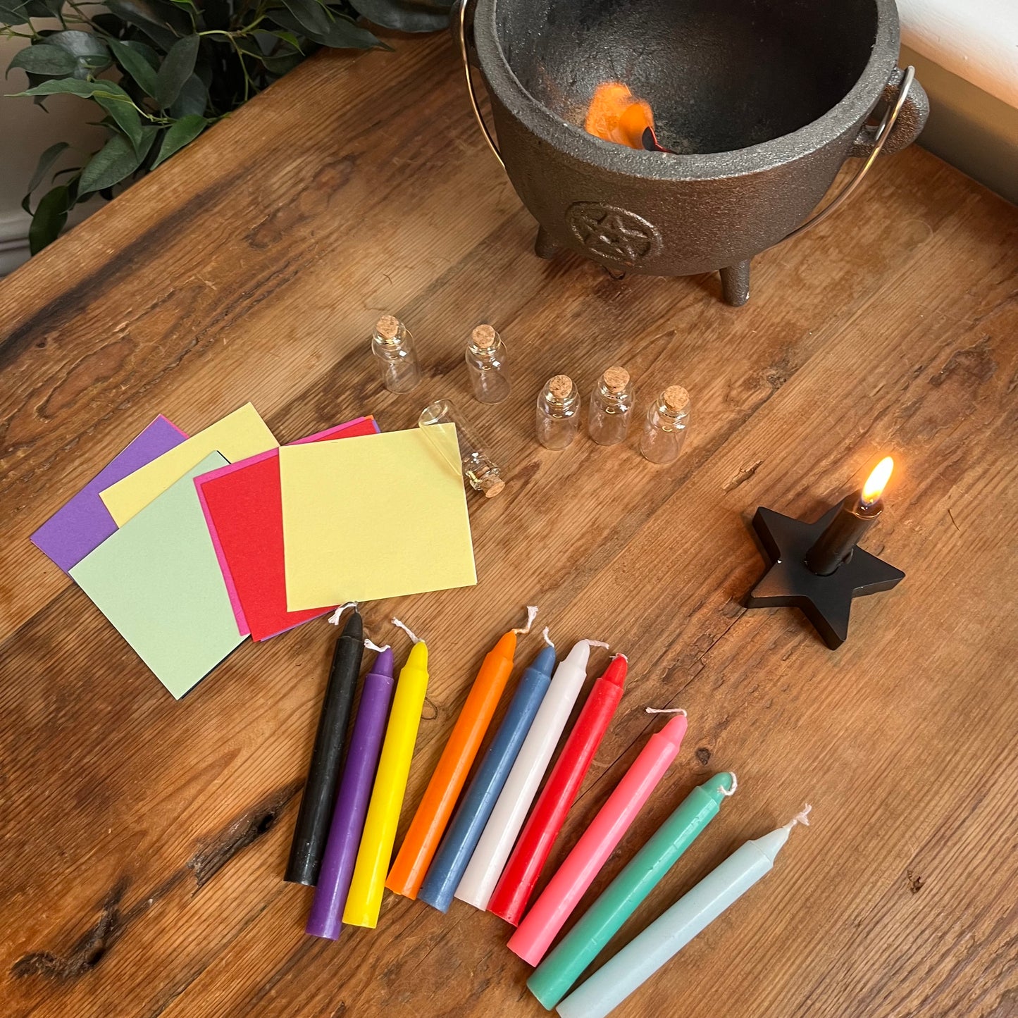 Displayed on a wooden table is the contents of the spell candle letterbox gift. 10 different coloured candles and coloured paper squares to match the candles, 6 small spell bottles to fill for your spells, a spell candle holder.