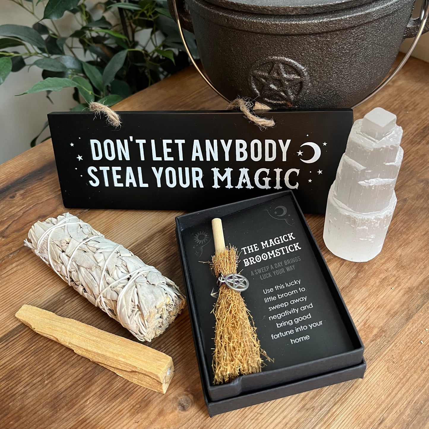 Contents of 'Protect your magic gift box'Pentagram mini broomstick in a black box, Selenite crystal mountain, Palo Santo stick, white sage smudge bundle, black wall sign with the words 'don't let anyone steal your magic'. pictured on a wooden table with a witches cauldron and green plant in the background.