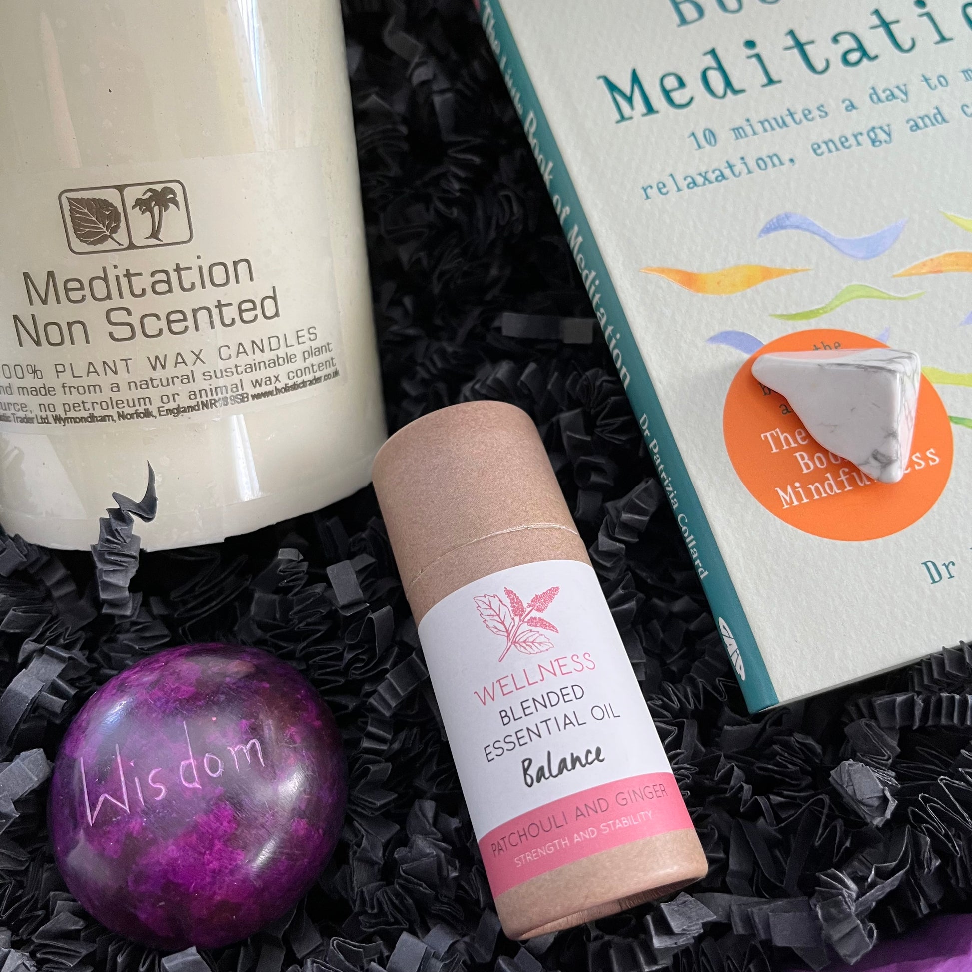 A wellness gift set arranged in black shredded paper, featuring a white plant wax candle jar labeled 'Meditation Non Scented,' a pink and white essential oil bottle labeled 'Wellness Blended Essential Oil Balance,' a purple marbled stone engraved with 'Wisdom,' and a book titled 'Buddhism and Meditation.'.