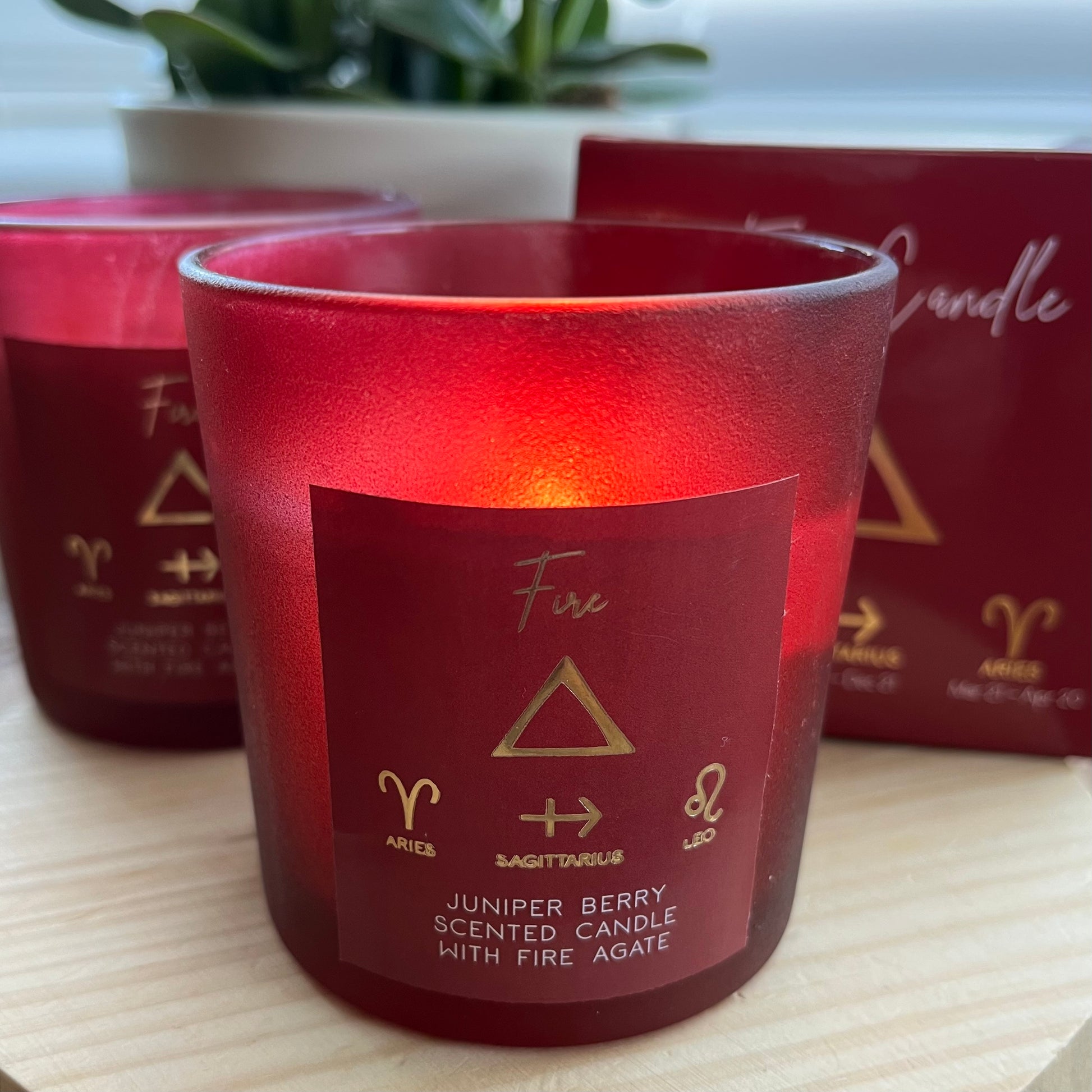 'Fire Element Candle' A red glass candle featuring the word fire and the symbol for the element of fire plus the symbols for the firey zodiac signs of Aries, Sagittarius and Leo. The candle is lit and gives off a warm glow. Alongside is its peresntation box with features the same words and symbols.