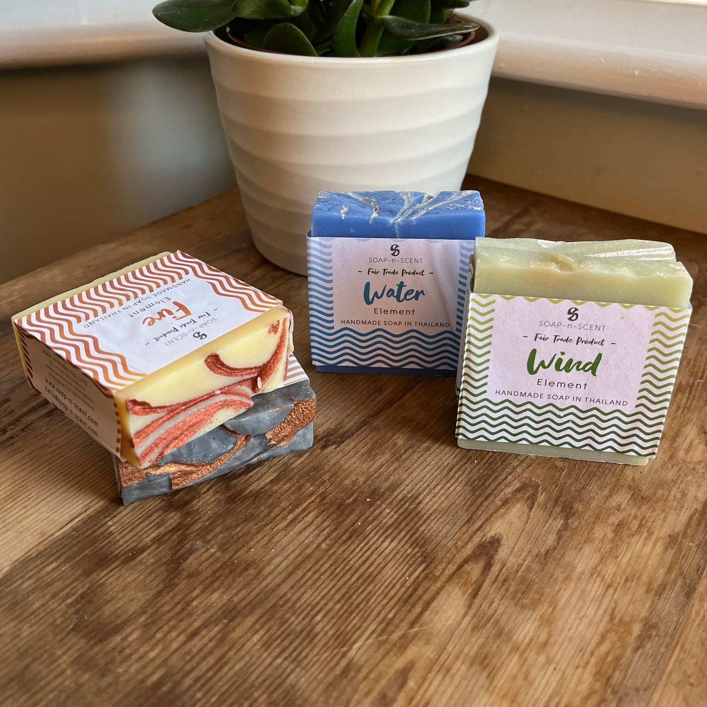 On a wooden table sits the contents of our 'Elements soap collection letterbox gift' 4 bars of soap named after the elements of wind, air, fire and water. wind is green with white pattern, earth is charcoal with bronze pattern, fire is cream with rust pattern, water is blue with white pattern. A green plant can be seen in the background.