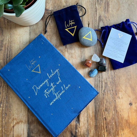 The contents of our 'Water signs elemental gift box' - a blue velvet notebook featuring inspiring words and the symbol for water. A grey stone decorated with the elemental symbol for water alongside its matching blue velvet pouch also featuring the same symbol, 5 tumble stones representing water Lapis Lazuli, Black Obsidian, clear Quart, Amethyst and Red Jasper and their pouch and information card. Photographed on a wooden table with a plant in the background.