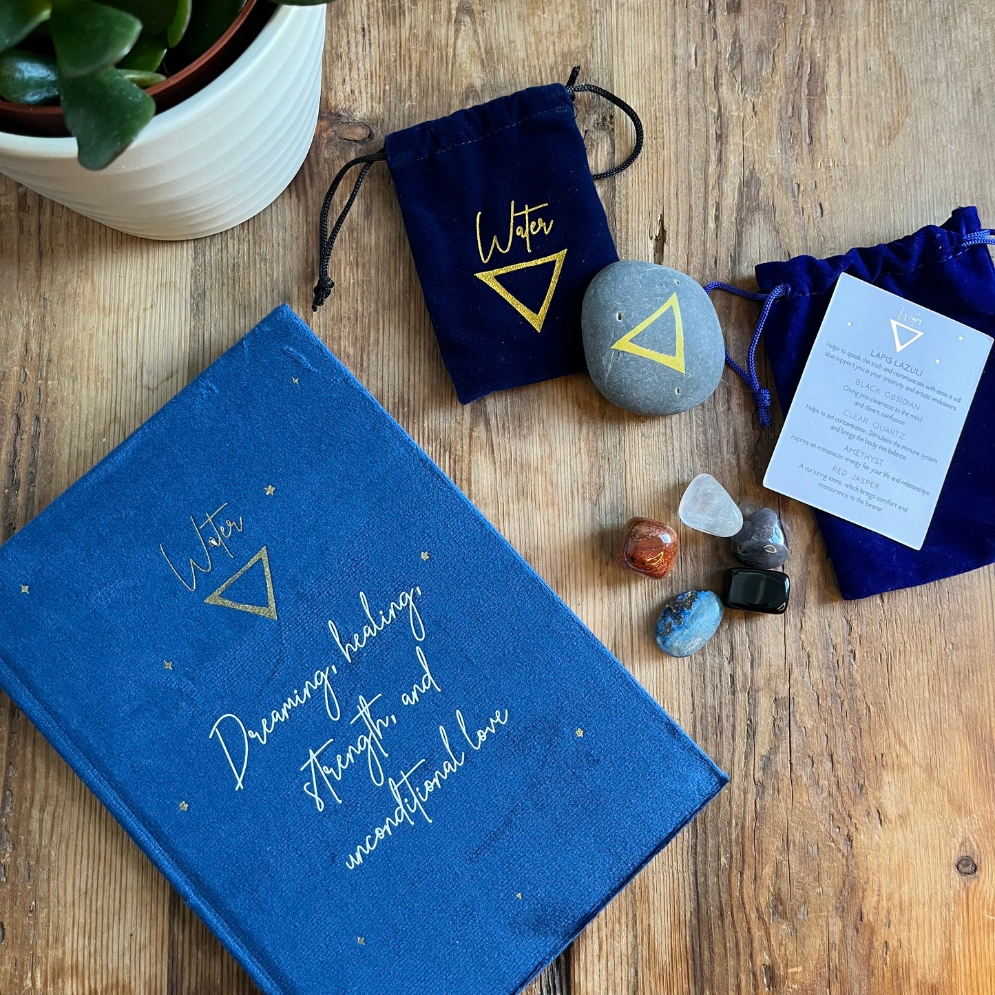 The contents of our 'Water signs elemental gift box' - a blue velvet notebook featuring inspiring words and the symbol for water. A grey stone decorated with the elemental symbol for water alongside its matching blue velvet pouch also featuring the same symbol, 5 tumble stones representing water Lapis Lazuli, Black Obsidian, clear Quart, Amethyst and Red Jasper and their pouch and information card. Photographed on a wooden table with a plant in the background.