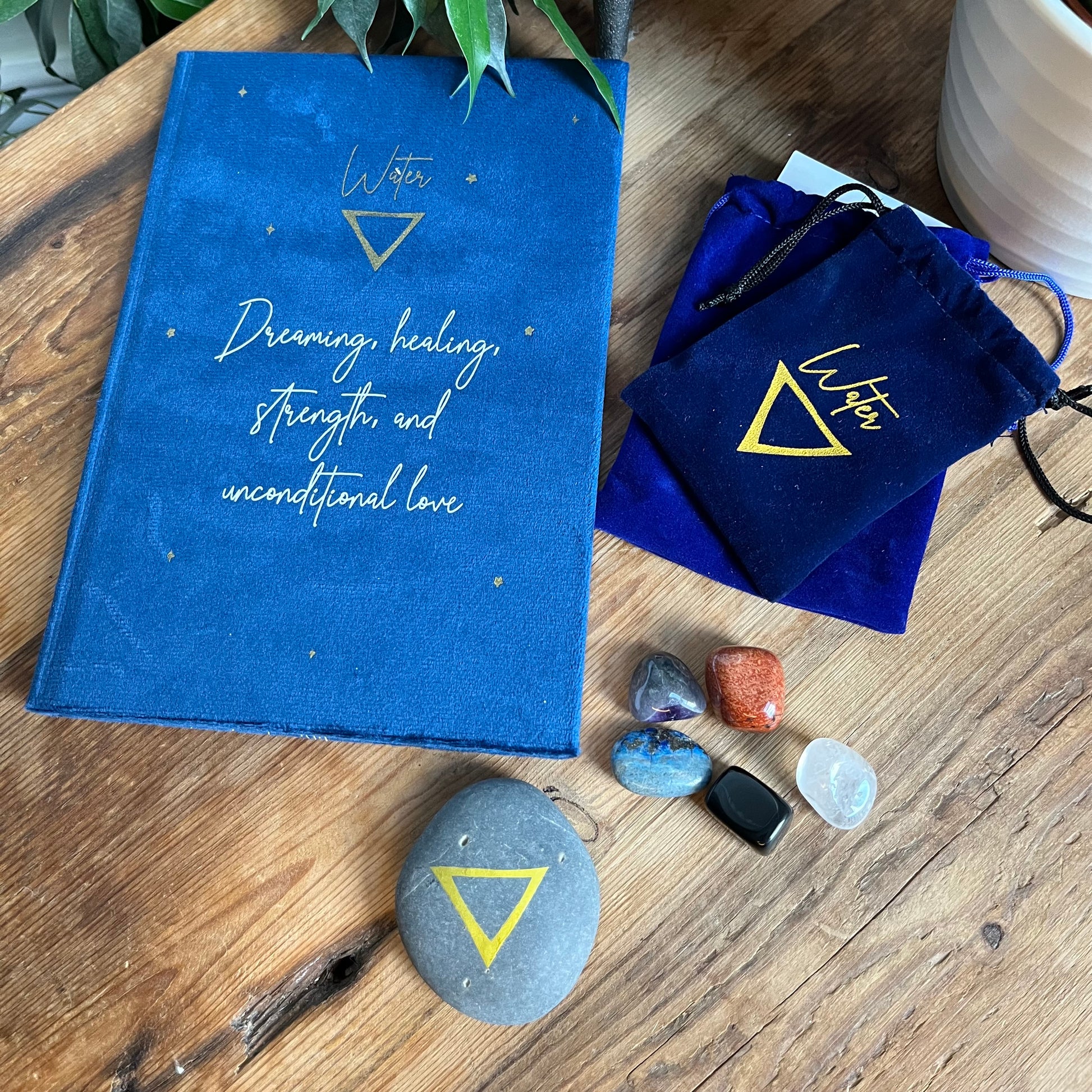 The contents of our 'Water signs elemental gift box' - a blue velvet notebook featuring inspiring words and the symbol for water. A grey stone decorated with the elemental symbol for water alongside its matching blue velvet pouch also featuring the same symbol, 5 tumble stones representing water Lapis Lazuli, Black Obsidian, clear Quart, Amethyst and Red Jasper and their pouch and information card. Photographed on a wooden table with a plant in the background.