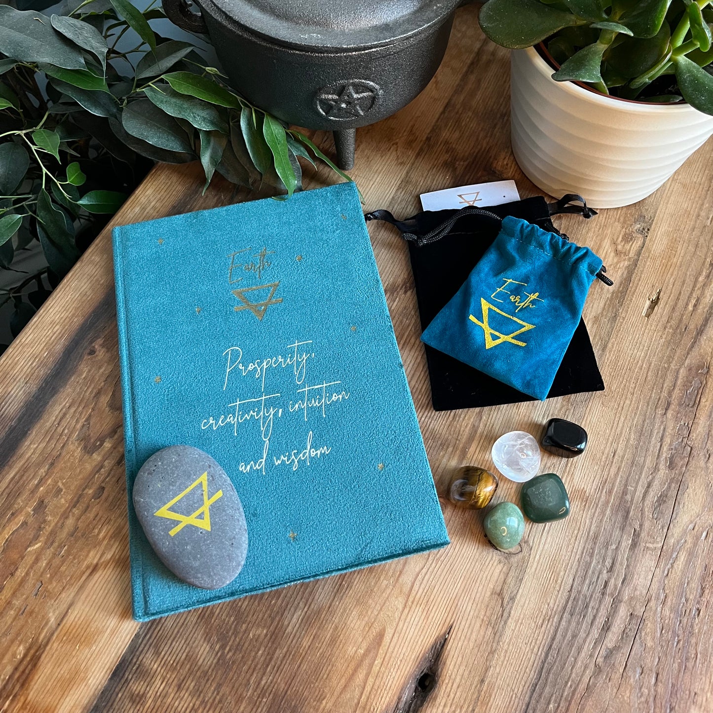 Contents of our 'Earth sign elemental gift box' - a Green velvet notebook featuring the elemental symbol for earth and inspiring words, two velvet pouches 1 containing a stone which features the elemental symbol for earth and the other contains tumbled stones associated with the element of earth. Displayed on a wooden table with a plant in the background 