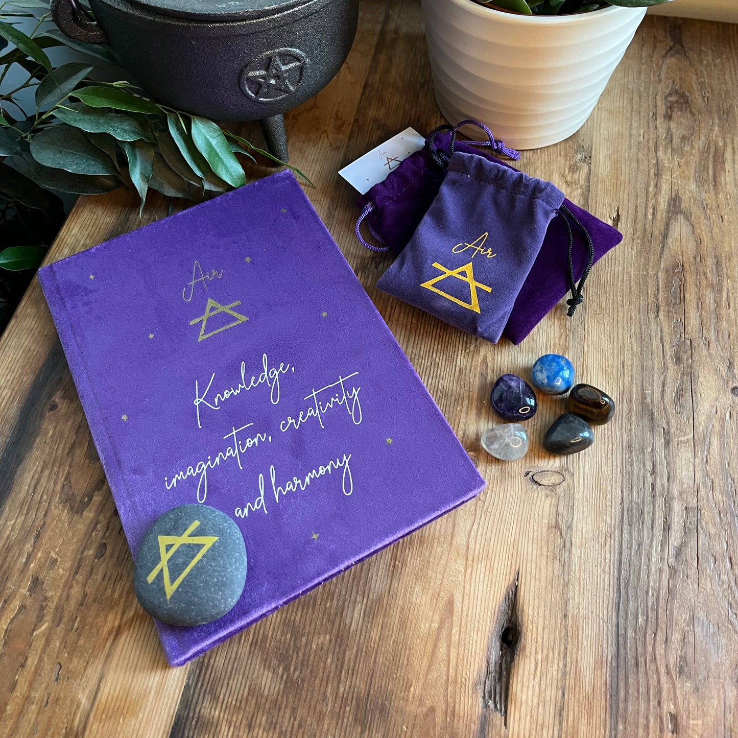 On a wooden table sits the contents of our 'Air Element Gift Box' a purple velvet notebook with inspiring words and the word air and the symbol for air. A grey rune stone decorated with the symbol for air alongside its purple velvet pouch also featuring the symbol for air. 5 tumble stones Lapis Lazuli, Amethyst, tigers eye, fluorite and labradorite, representing the element of air sit alongside their purple velvet pouch. In the back ground is a plant.