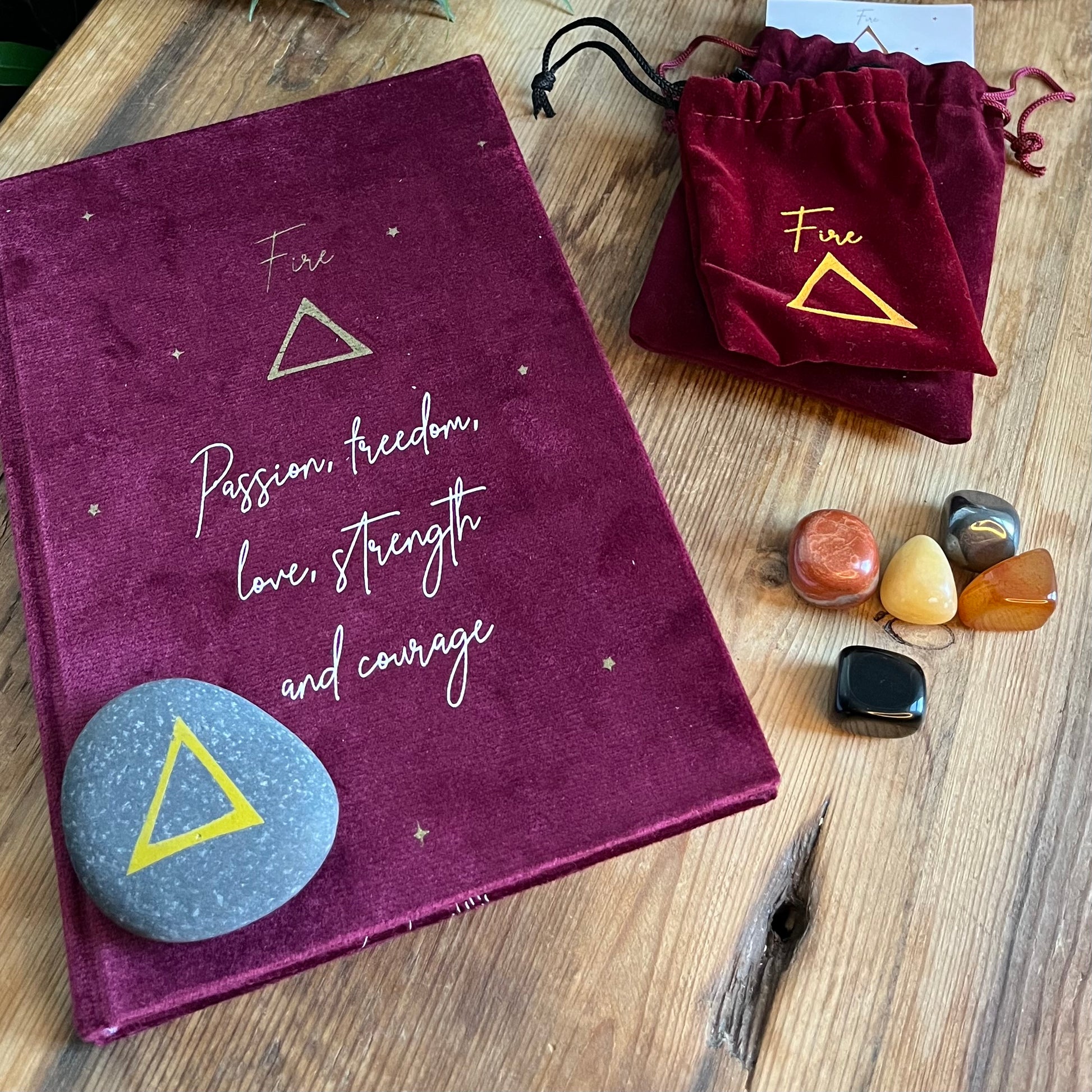 The contents of our 'Fire Element Gift Box' are shown on a wooden table. We can see a red velvet notebook with the word fire on the cover with the symbol for fire and inspirational words. A grey stone with the symbol for fire inscribed onto it. 5 tumblestones connected to the fire element Black Obsidian, yellow Aventurine, Carnelian, Hematite and red Jasper also shows 2 red velvet pouches one for the rune the other for the tumblestones.