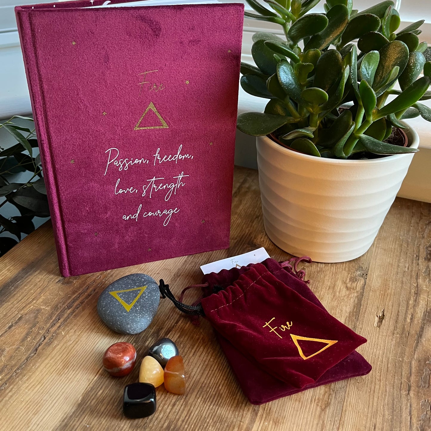 The contents of our 'Fire Element Gift Box' are shown on a wooden table. We can see a red velvet notebook with the word fire on the cover with the symbol for fire and inspirational words. A grey stone with the symbol for fire inscribed onto it. 5 tumblestones connected to the fire element Black Obsidian, yellow Aventurine, Carnelian, Hematite and red Jasper also shows 2 red velvet pouches one for the rune the other for the tumblestones.