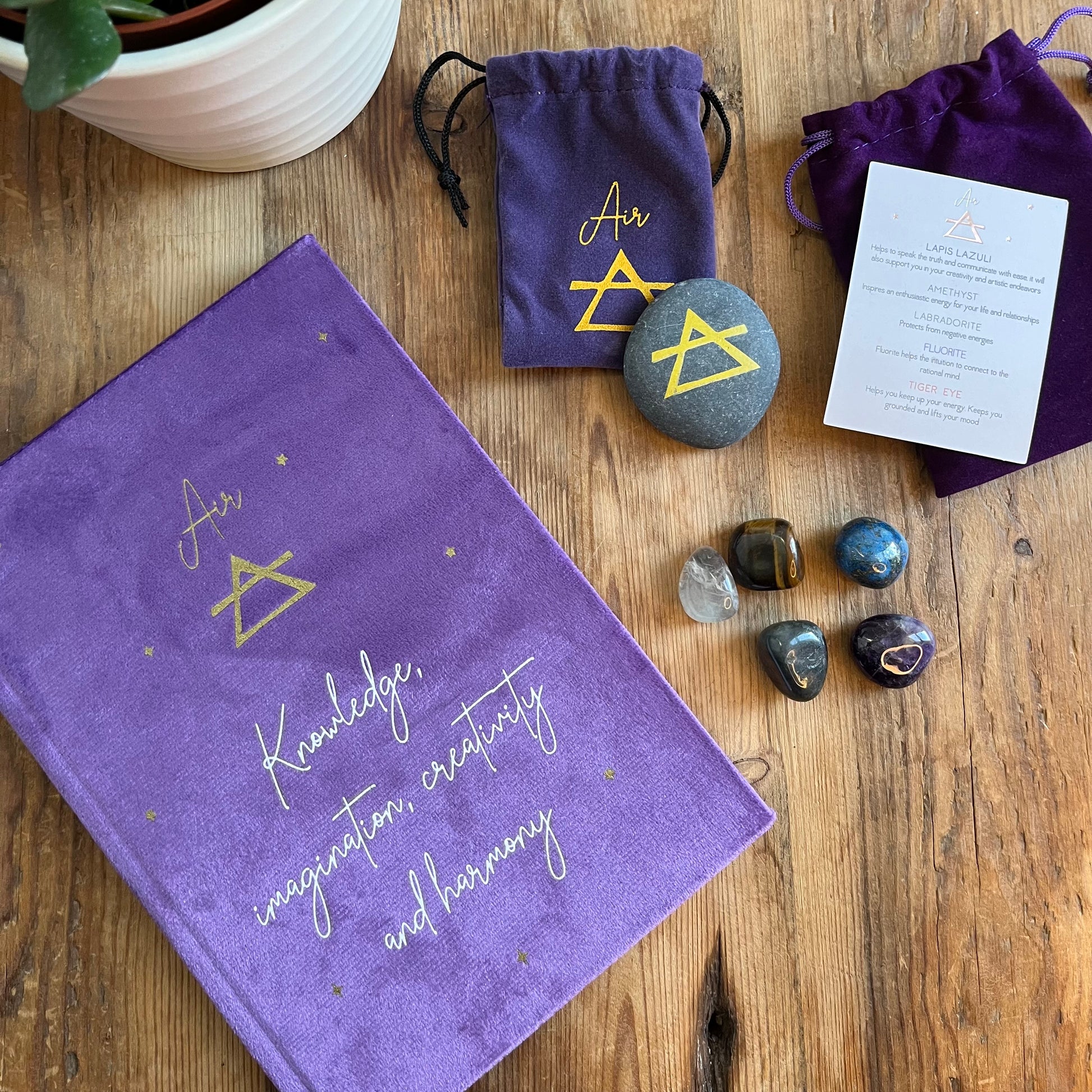 On a wooden table sits the contents of our 'Air Element Gift Box' a purple velvet notebook with inspiring words and the word air and the symbol for air. A grey rune stone decorated with the symbol for air alongside its purple velvet pouch also featuring the symbol for air. 5 tumble stones Lapis Lazuli, Amethyst, tigers eye, fluorite and labradorite, representing the element of air sit alongside their purple velvet pouch. In the back ground is a plant.