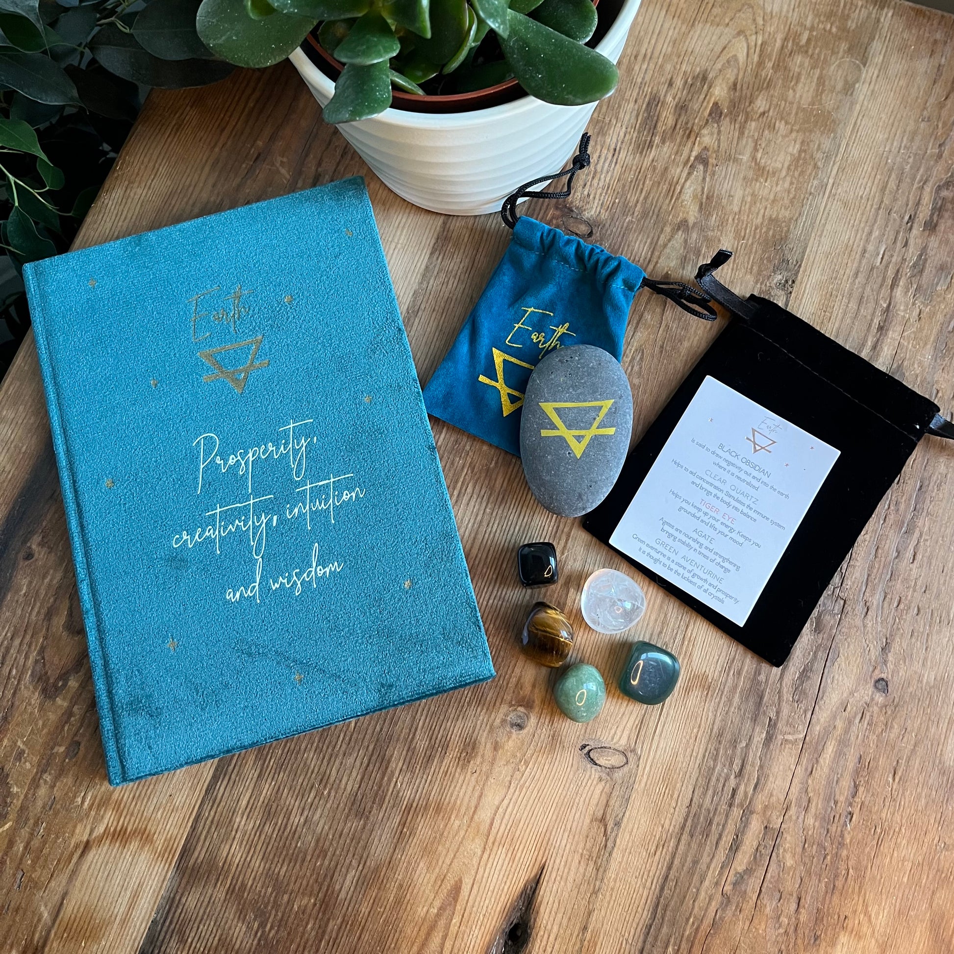 The contents of our 'Earth  sign Elemental Gift Box'  are shown on a wooden table, we can see a Green velvet notebook featuring the word Earth and the symbol for Earth and inspiring words, 2 velvet pouches, containing earth element rune and featuring the word earth and a symbol for earth, one  containing earth element tumblestones .