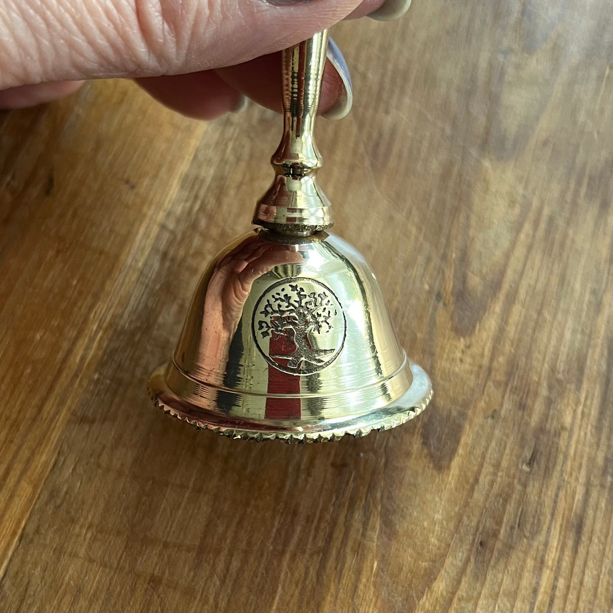 A small polished brass bell with a Tree of Life emblem, held by a hand over a wooden tabletop.