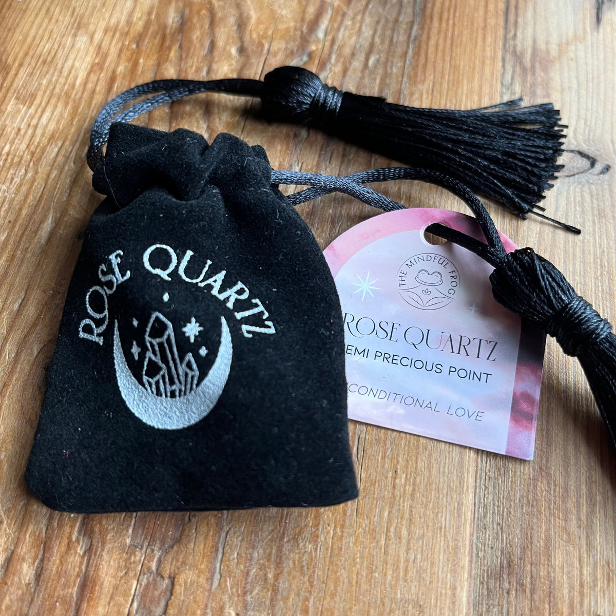 A small black velvet drawstring pouch with a silver-toned rose quartz crystal design, shown on a wooden surface next to a pink and white product tag labeled 'Rose Quartz Semi Precious Point'.