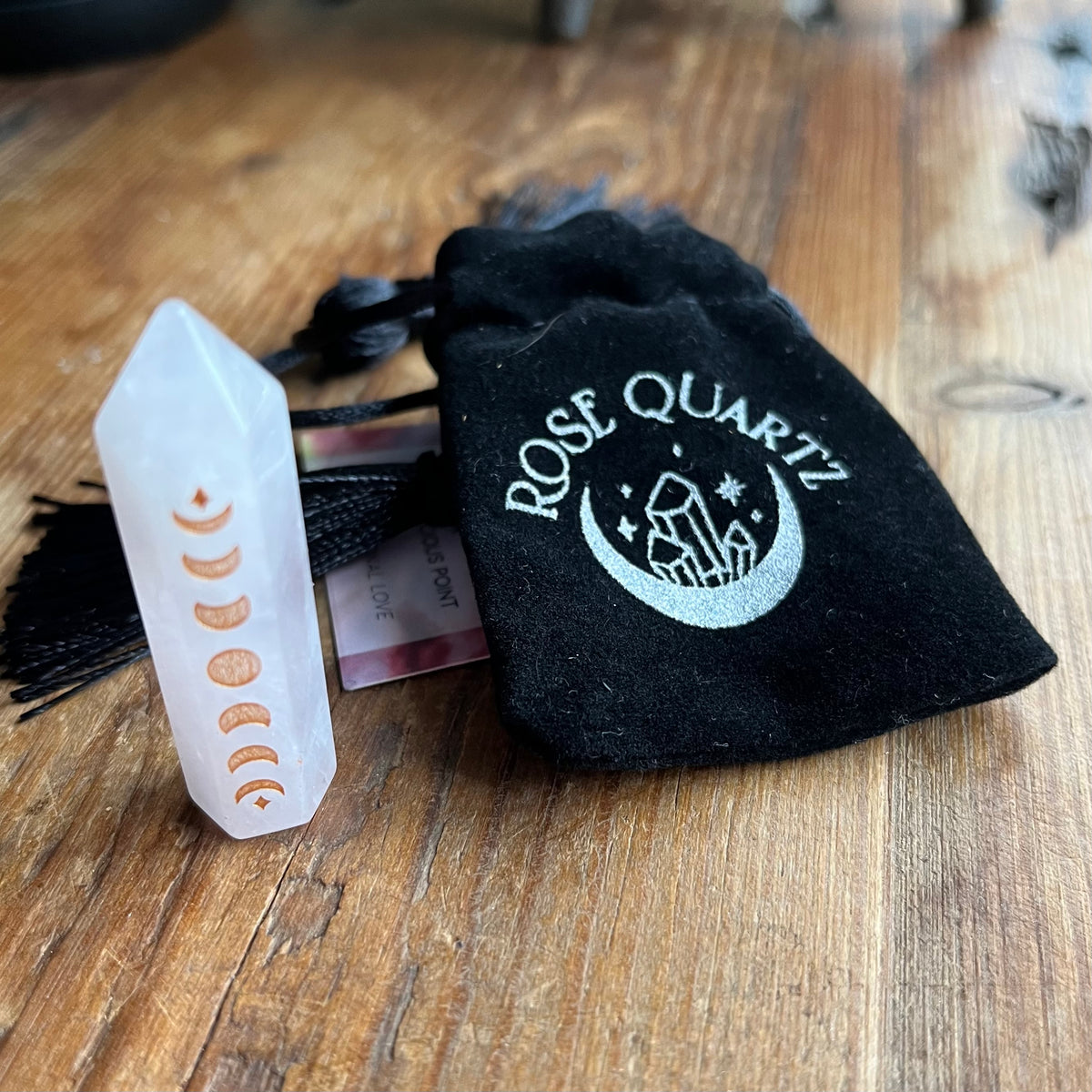 A rose quartz crystal tower showing the moon phase alignment, displayed next to a black velvet drawstring pouch embroidered with 'ROSE QUARTZ' and a crescent moon design, on a wooden tabletop.
