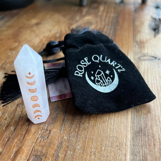 A rose quartz crystal tower showing the moon phase alignment, displayed next to a black velvet drawstring pouch embroidered with 'ROSE QUARTZ' and a crescent moon design, on a wooden tabletop.
