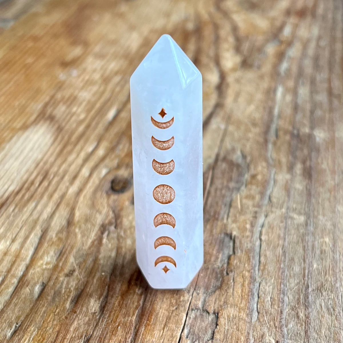 A pointed Pink crystal tower with engraved copper-colored moon phases, standing upright on a rustic wooden surface.