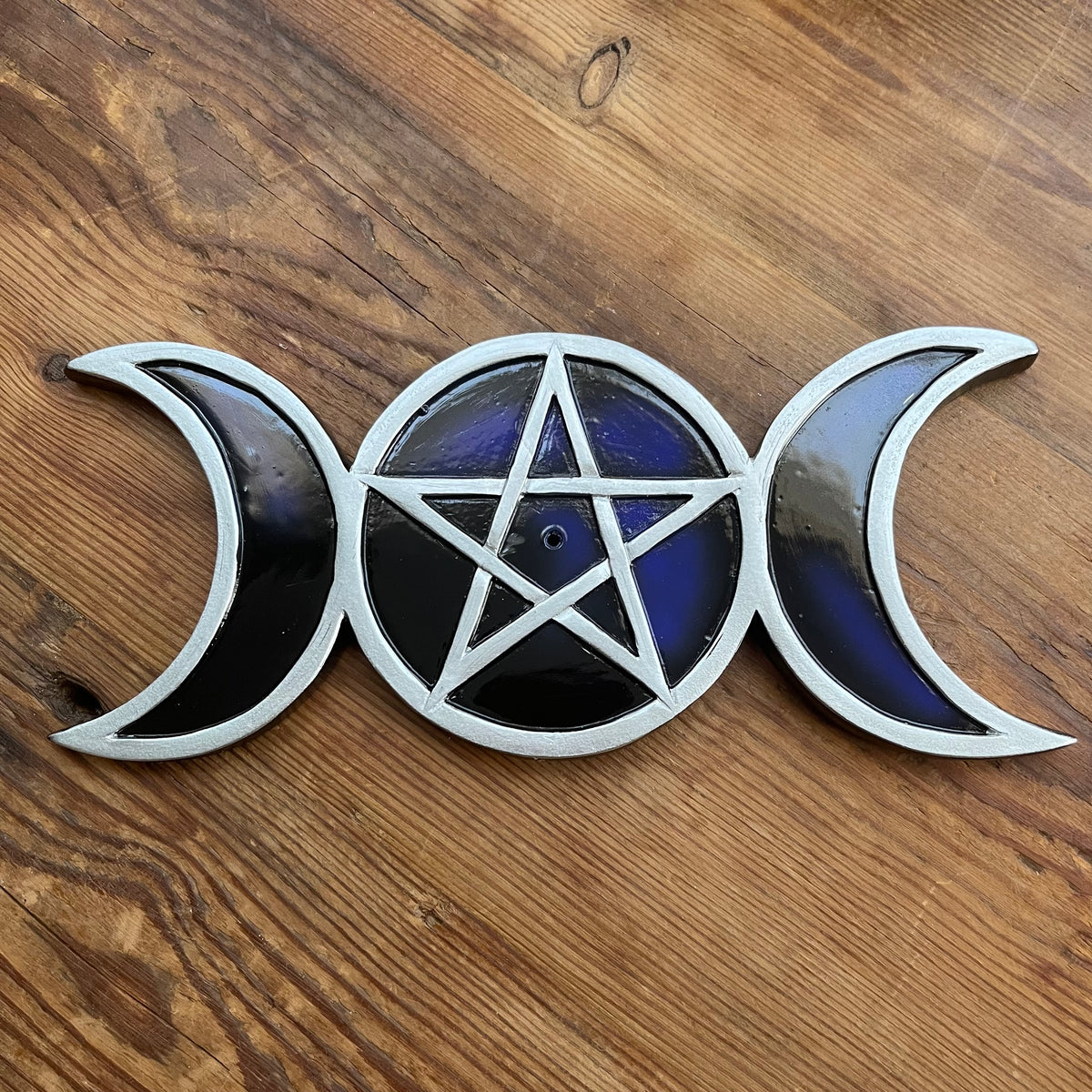 A silver-toned resin + metal incense holder featuring a triple moon and triple star pentagram design, with dark blue and black enamel details, resting on a wooden surface.